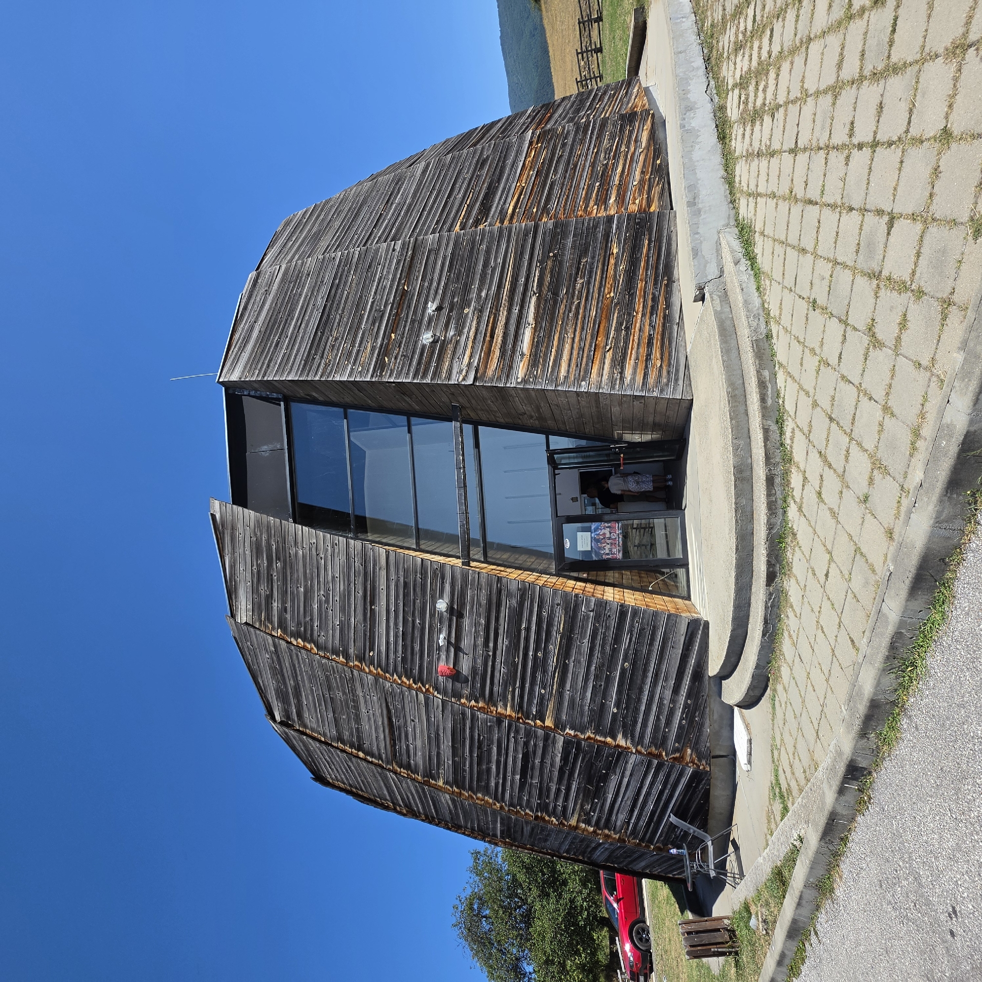 In Dorkovo, Bulgaria, a distinctive, conical wooden building stands against a clear blue sky. The structure is clad in dark, weathered horizontal wooden planks, with some showing a reddish-brown undertone. A large, angled glass facade is set into the front of the building, revealing an entrance with glass doors. Two individuals are visible standing just inside the entrance. To the left of the building, a bright red car is parked on a grassy area next to a tree. A small table with two chairs is also situated near the building. The ground in front of the entrance is paved with square concrete tiles, and a concrete walkway leads to the building's entrance. In the background, a grassy hillside slopes upward, with a wooden fence visible along its crest. The scene is bathed in bright sunlight.