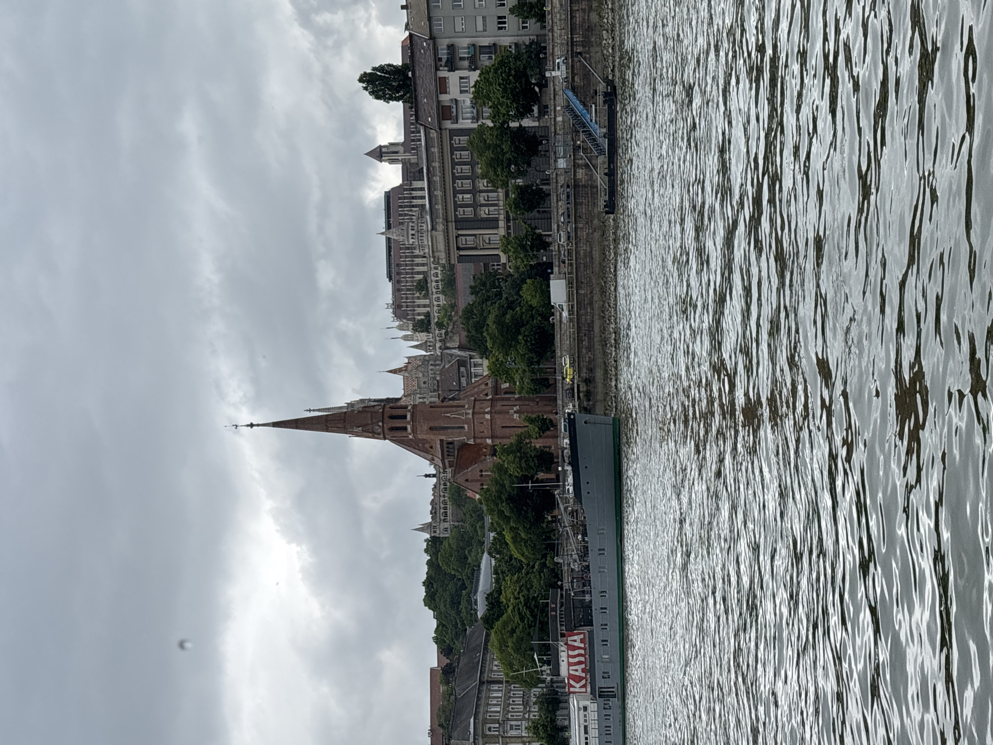 The image shows the Danube River in Budapest, Hungary. The river occupies most of the right half of the image, with visible ripples on its surface. On the left side of the image, a cityscape stretches along the riverbank. Buildings of varying heights and architectural styles are visible, some with trees lining the riverfront. Prominently featured is a tall, brick-colored church with a distinctive spire. A boat with the word "KASSA" on its side is docked along the riverbank. The sky is overcast, with grey clouds filling the upper portion of the image.