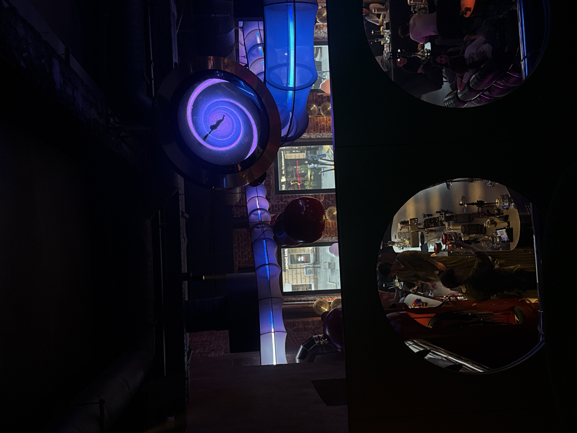 The image shows a night view of a bar or restaurant in Moscow, Russia. The view is partially obstructed by a dark, circular frame with two round openings. Through these openings, the interior of the venue is visible. The upper opening shows people sitting at a bar, while the lower opening displays a view of a counter with bar equipment.
To the left of the openings, a futuristic-looking, vertically arranged structure consists of a round screen with a purple swirling pattern, illuminated white and blue cylindrical sections, and metallic pipes. In the background, lit screens show scenes of the city outside.