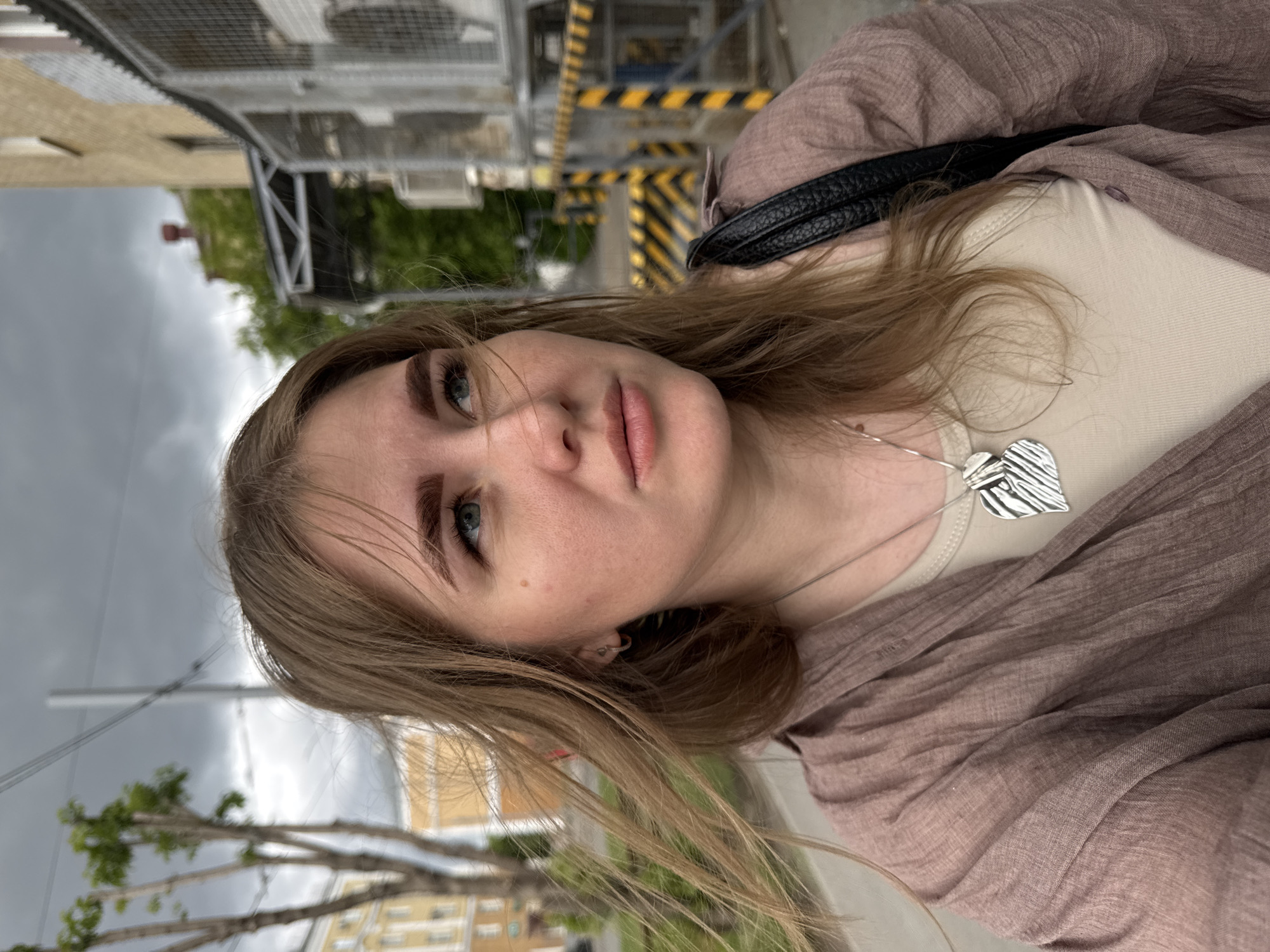 A selfie taken by a fair-skinned woman with blonde hair. She wears a beige top and a mauve-colored scarf, along with a silver leaf-shaped necklace. A black handbag strap is visible over her shoulder. The background shows elements of Perm, Russia, including a building with yellow and white trim, and gray overcast skies. A metal structure with black and yellow hazard stripes is also visible.