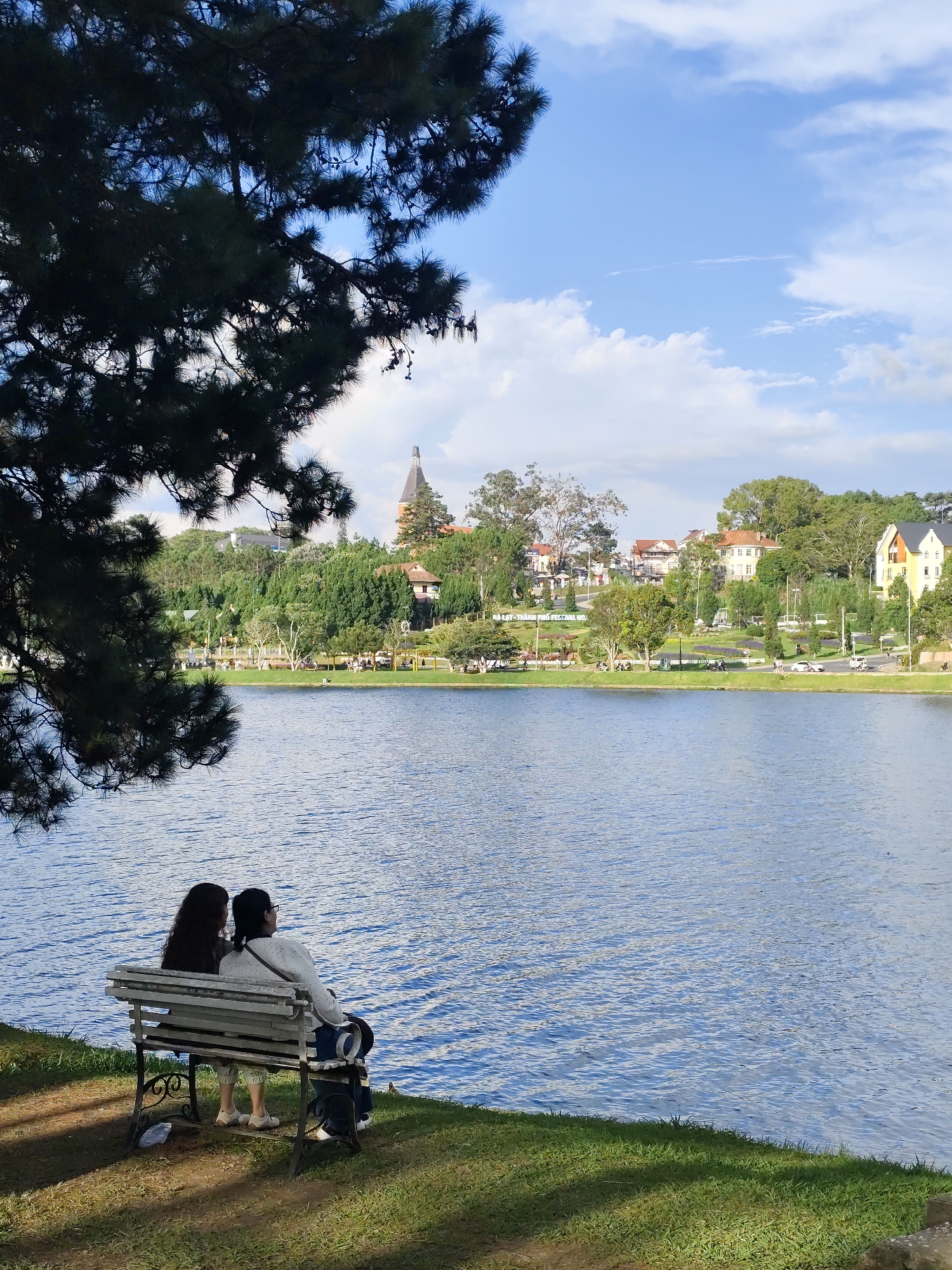 Two individuals are seated on a weathered park bench on a grassy incline overlooking a tranquil body of water. The person on the left has long, dark, wavy hair and is wearing a dark shirt. The person on the right has dark hair, wears glasses, and is attired in a white sweater and light-colored pants. They are both facing the water, seemingly engaged in conversation or contemplation. A dark-colored backpack rests on the bench beside the person on the right. The lake's surface exhibits gentle ripples, reflecting the bright, partly cloudy sky. Across the water, a vibrant cityscape unfolds, characterized by lush greenery, a variety of architectural styles including houses and buildings with distinctive roofs, and the presence of trees lining the shore. In the distance, a prominent tower with a dark, conical roof stands out against the skyline. The scene is illuminated by natural daylight, suggesting it was captured during daytime hours. This scene depicts a serene moment at Xuan Huong Lake in Da Lat, Vietnam, a popular tourist destination known for its picturesque landscapes and French colonial architecture.