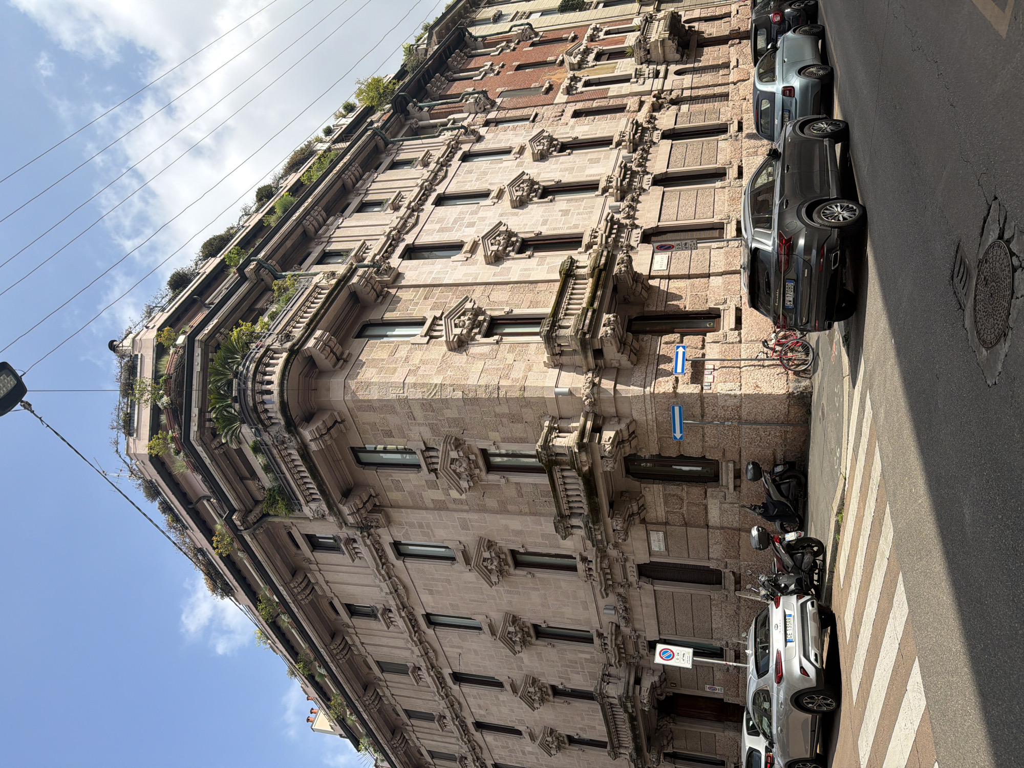 A low-angle, exterior shot depicts a building with ornate architectural details and greenery on its upper levels. The structure is predominantly light brown, with recurring geometric patterns around the windows. Parked along the street are cars and motorcycles, with a zebra crossing visible in the foreground. Street signs are mounted on the building's facade. The image is taken in Milan, Italy.