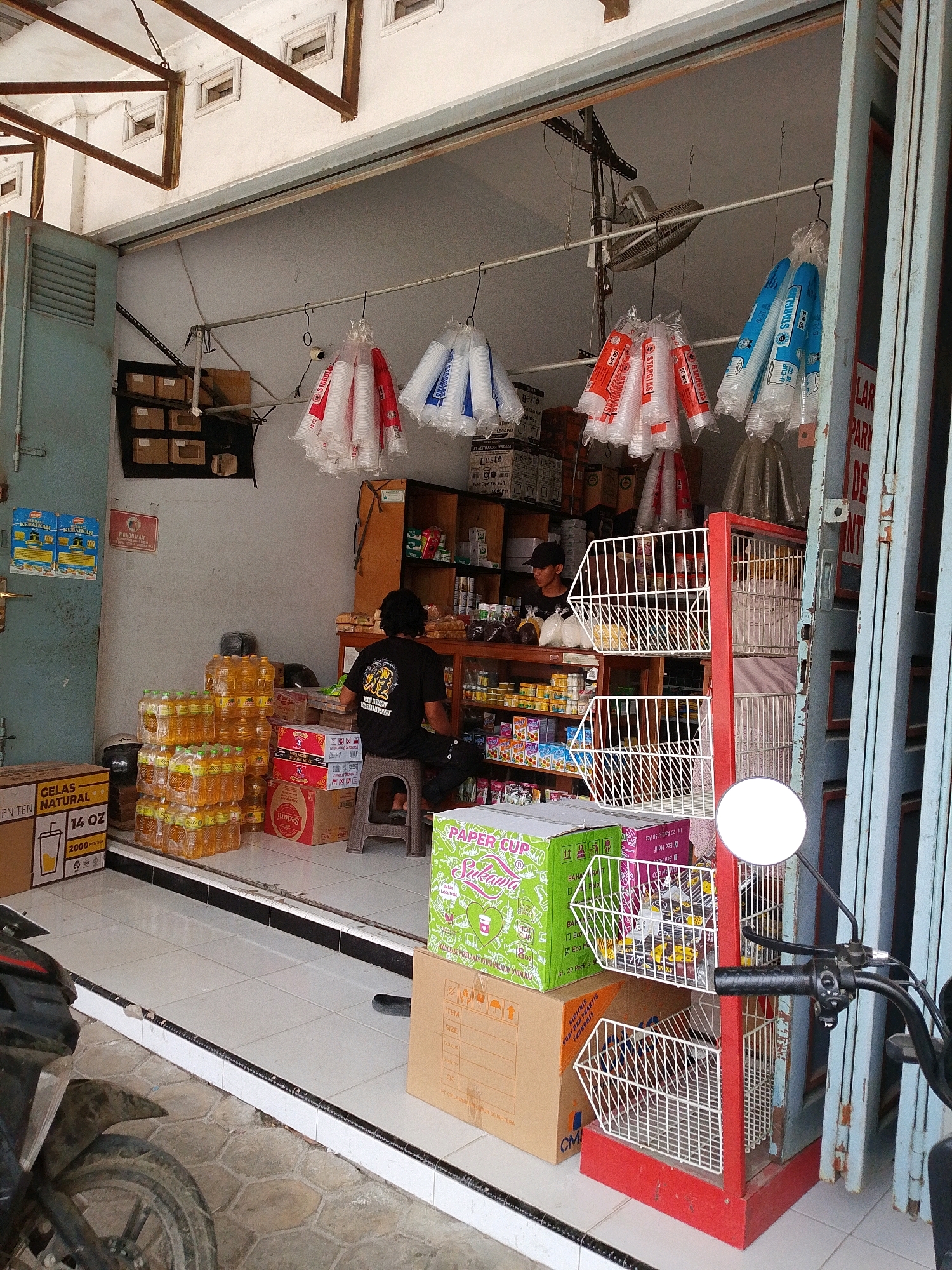 The image depicts the exterior of a small shop located in Grogol, Indonesia. The shop's entrance is open, revealing an interior filled with various goods. In the foreground, stacks of bottled cooking oil and cardboard boxes are visible on the tiled floor. A motorcycle is parked to the left.
Inside the shop, a person wearing a black t-shirt and cap is seated on a stool, facing away from the camera and attending to some items. Shelves are lined with products, and several plastic cups are hanging from the ceiling. A red display rack with wire baskets is positioned near the entrance. Beyond the immediate shop area, a closed metal gate is visible on the right side of the frame. The overall impression is that of a local convenience store or small grocery outlet.