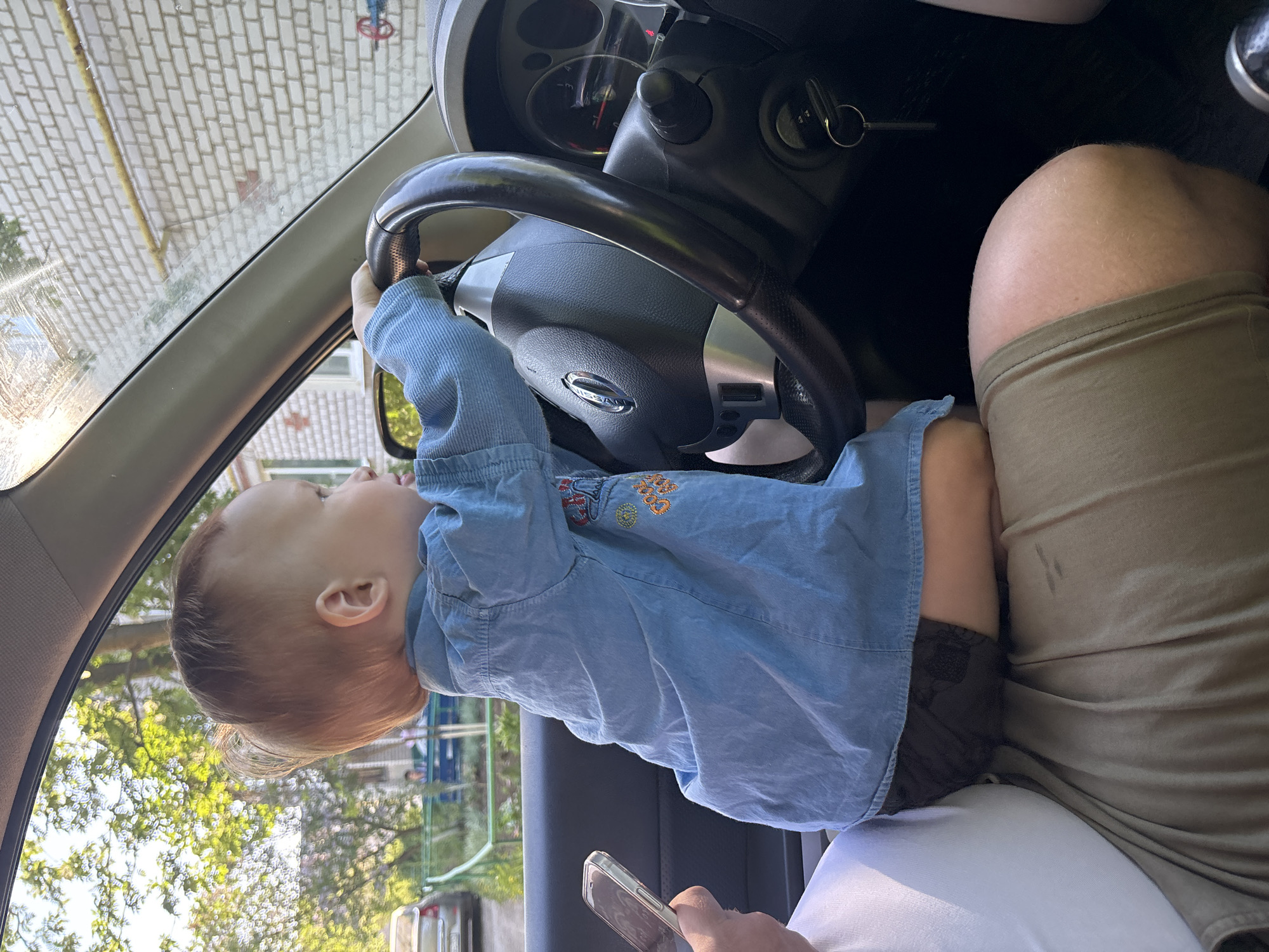 A toddler sits on the lap of an adult in the driver's seat of a car, holding the steering wheel. The car is likely stationary, as indicated by the background view of a residential street and the presence of the key in the ignition. The adult is holding a smartphone. The photograph appears to have been taken in the city of Горный, Russia.