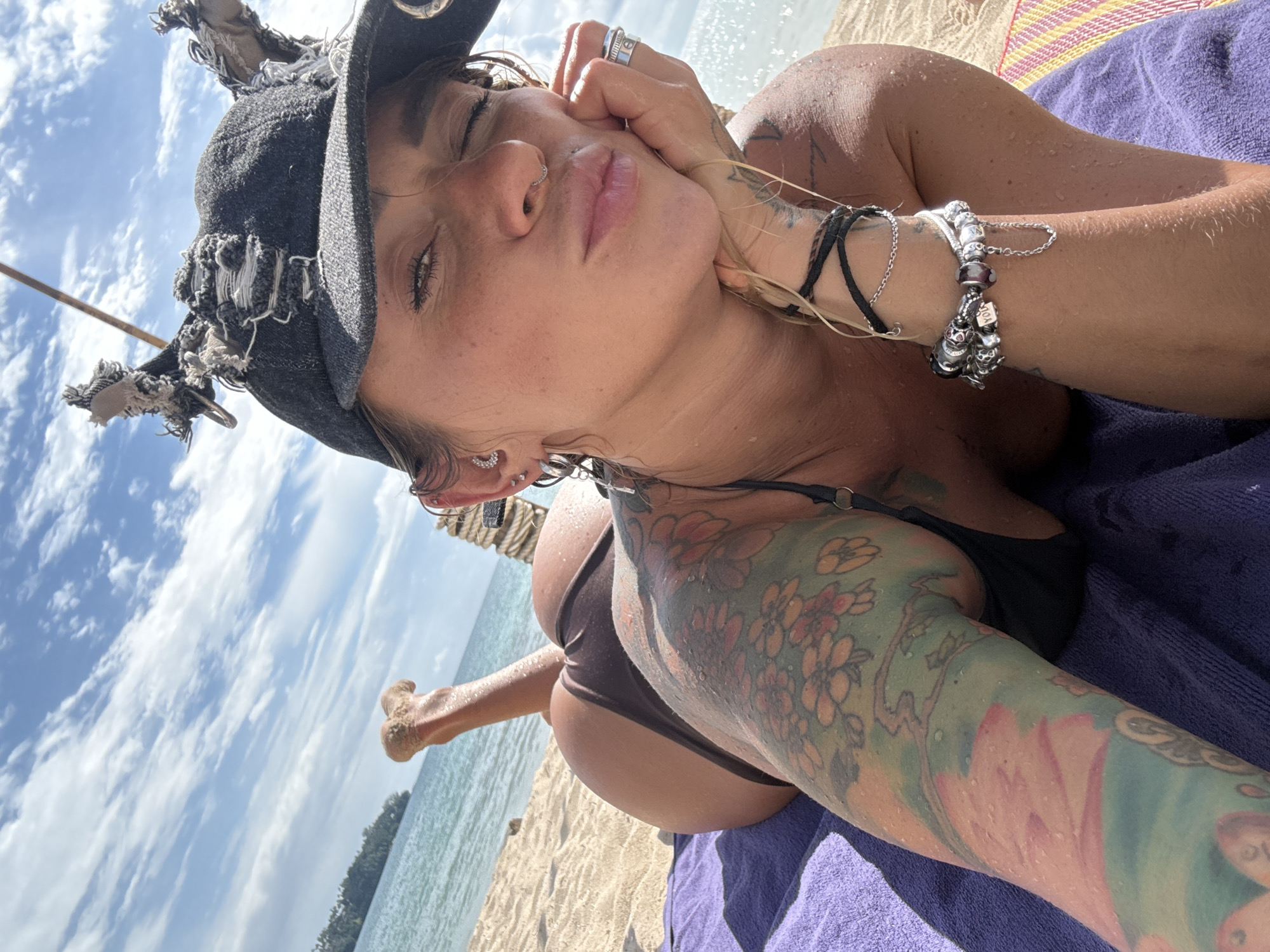 A person lies on their side on a beach in Choeng Thale, Thailand, possibly sunbathing or relaxing. The person is wearing a dark bikini and a distressed black baseball cap. Their skin appears wet, with water droplets visible. The person has several tattoos on their left arm and shoulder, including floral designs and other imagery. They are also wearing jewelry on their wrists and a ring on their right hand.
The scene is a sandy beach with the ocean in the background. The sky is blue with some scattered clouds, suggesting it is daytime, likely sunny. The person is lying on a purple towel, which is placed on a striped mat. The overall impression is one of leisure and enjoyment of a tropical environment. The lighting suggests it is a bright, sunny day, and the water droplets on the skin indicate either recent swimming or spray from the ocean. No visible text is present in the image.