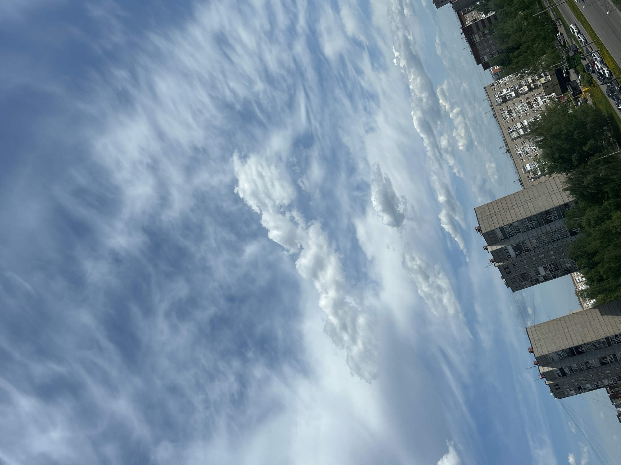 The image shows a view of the sky and buildings in Izhevsk, Russia. The sky is predominantly blue with wispy cirrus clouds stretching across the frame. There are also some cumulus clouds interspersed. Along the right edge of the image, a series of high-rise apartment buildings are visible, with trees partially obscuring some sections. A road and parked cars can be seen to the right of the buildings. The image is tilted at an angle, making the buildings appear to lean towards the left.