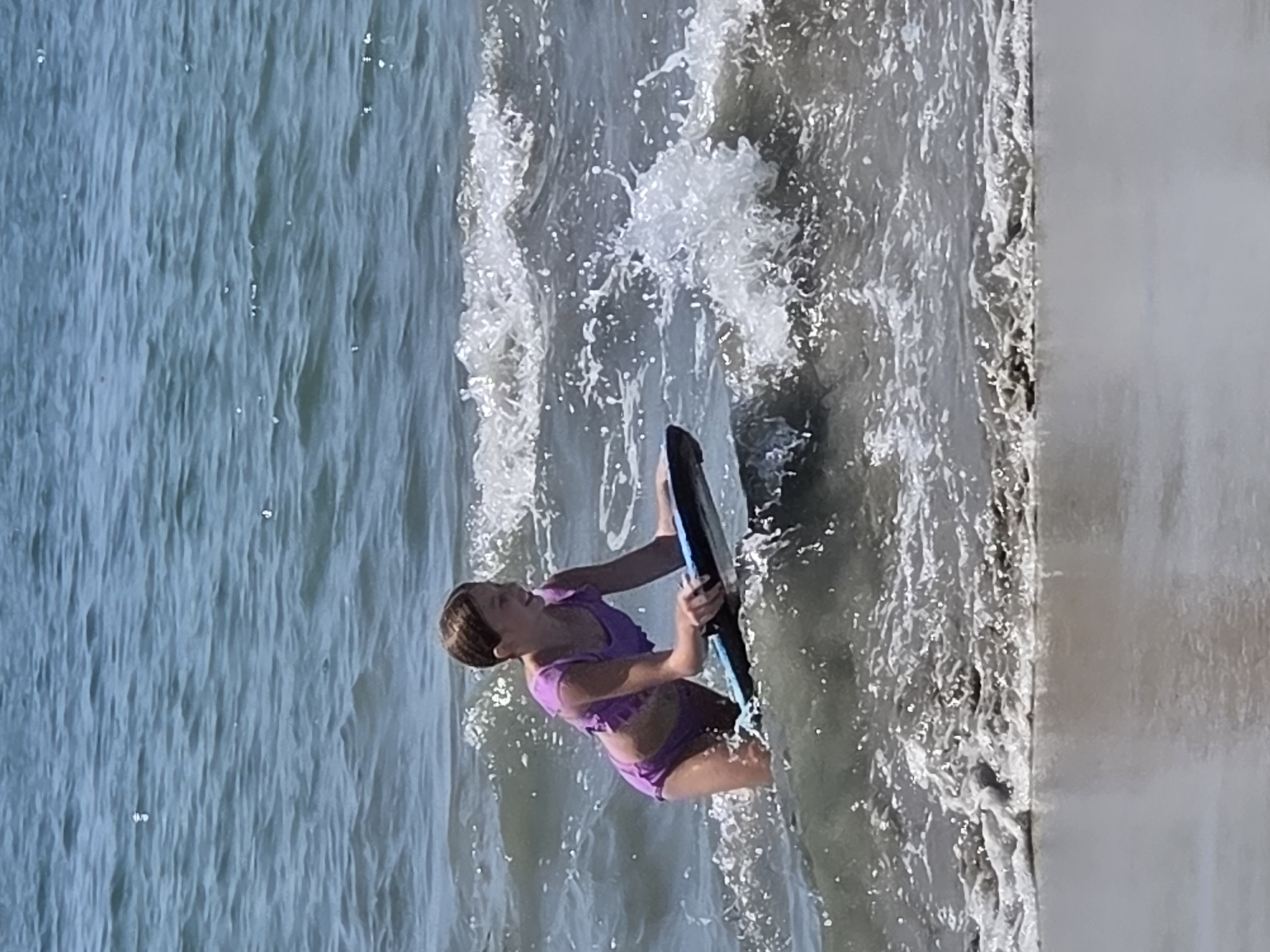 A young girl is bodyboarding in the ocean near a concrete structure. She is wearing a purple bikini and is positioned on the bodyboard with her arms holding onto it. The water is choppy and appears to be breaking around her. The ocean is a bluish-gray color, and there are whitecaps on the waves. The concrete structure is light gray and runs vertically along the right side of the frame. The scene is likely taking place during the day, given the lighting. The girl's facial expression is difficult to discern due to the angle and resolution of the image, but she appears to be engaged in the activity. There is no visible text in the image. The location context provided is Long Beach, United States of America.