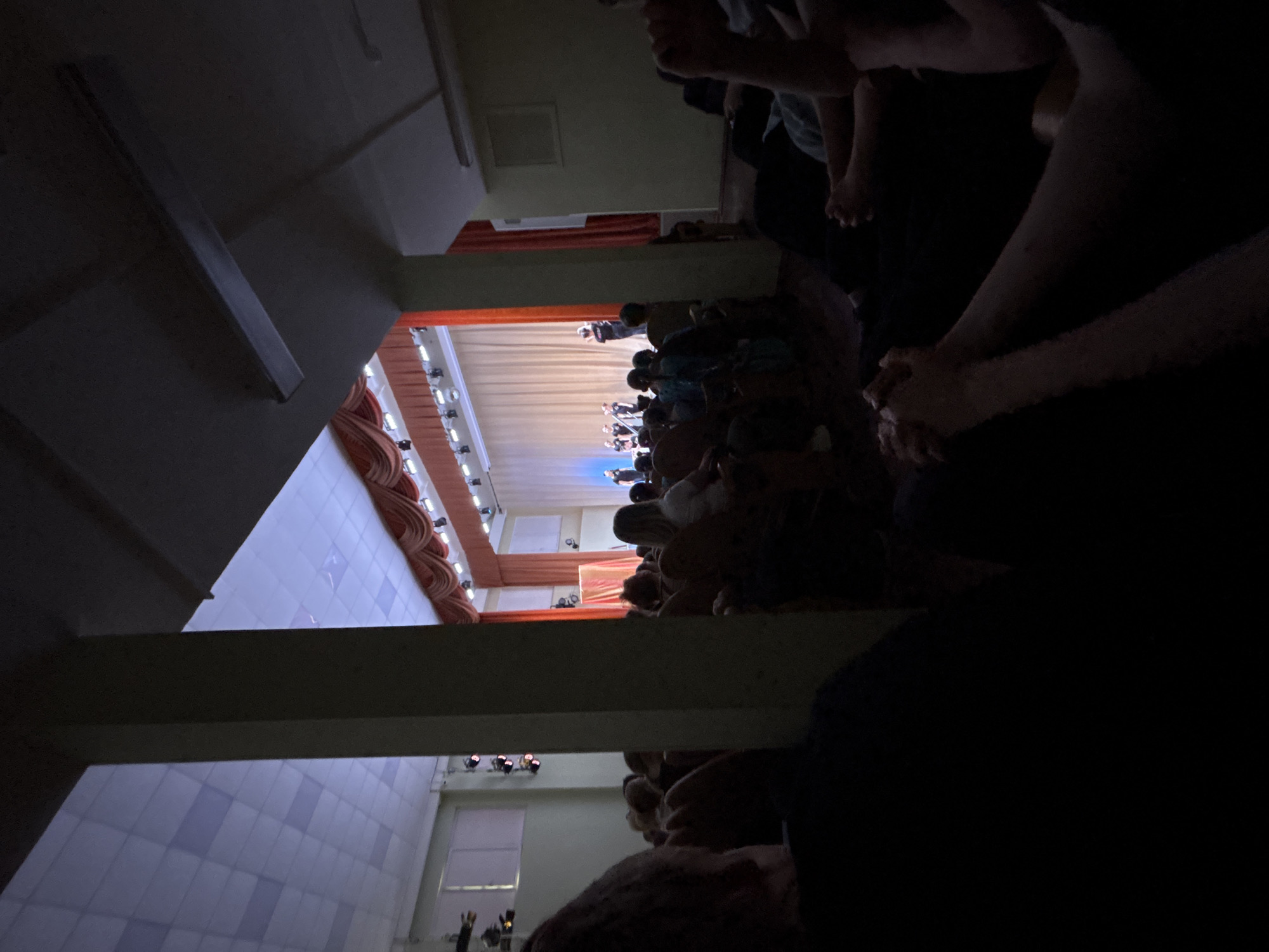 The image shows a view from an elevated position, possibly a balcony, inside what appears to be a theater or auditorium. Many people are seated below, facing a stage where a performance seems to be taking place. The stage is illuminated with lights, and the backdrop includes red fabric. The room has a tiled floor and a visible air conditioning vent. The image's perspective is slightly tilted. Given the mention of Saint Petersburg, Russia, this could be a theater in that city, such as the Mariinsky Theatre or Alexandrinsky Theatre. However, without additional details, a precise identification of the location is not possible.