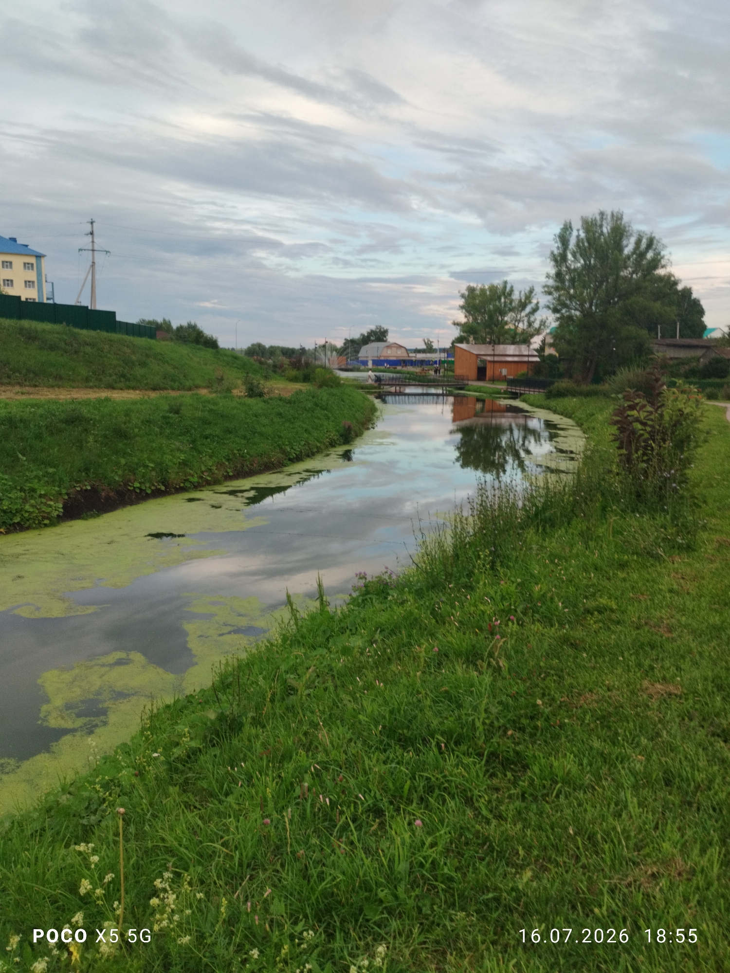 The image depicts a tranquil outdoor scene in Aksubayevo, Russia, featuring a narrow canal flowing through a grassy landscape under a cloudy sky. The canal's surface is partially covered with green algae and reflects the sky and surrounding foliage. On the left bank, a sloped grassy hill is visible, topped with a green fence and a building with a blue roof. Further in the background, several trees and structures, including a wooden building and a small bridge over the canal, can be observed. The right bank of the canal is lined with lush green grass and vegetation, with a pathway visible in the distance. The lower portion of the image displays a timestamp and camera model information.