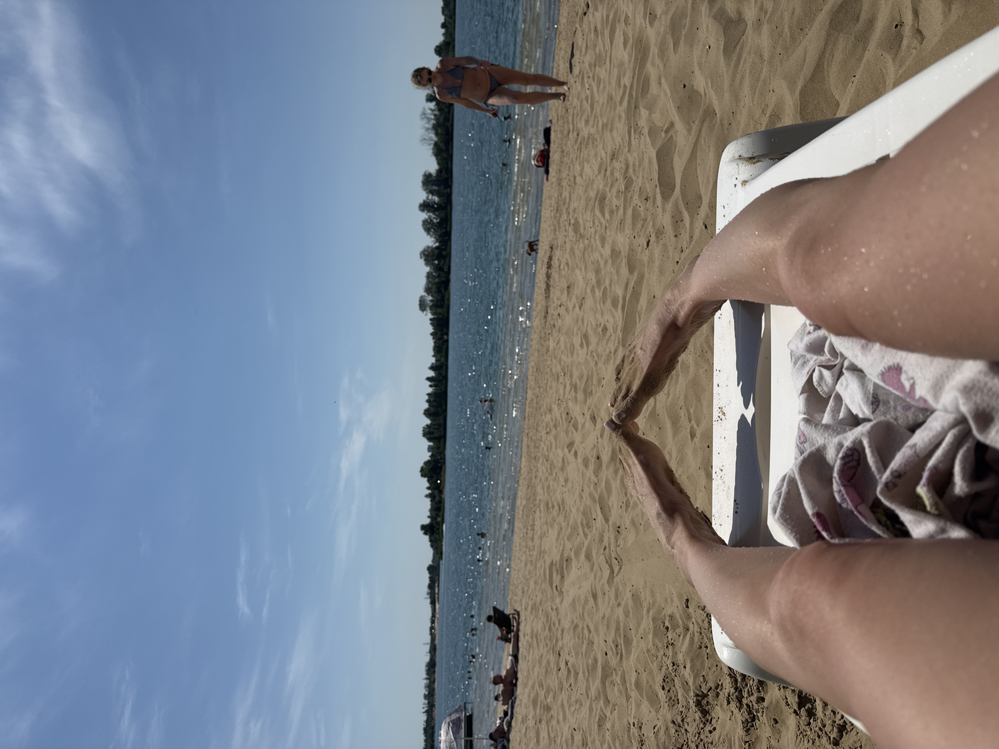 The image depicts a sunny day at a beach resort located on the shores of the Yelyzavetivka reservoir in Yelyzavetivka, Ukraine. The perspective is from someone lying on a white lounge chair, with their legs and feet prominently in the foreground. The sand is a light tan color, with visible footprints and textures. A towel with a floral pattern is draped over the lounge chair.
In the middle ground, the sandy beach slopes down to the shimmering blue water of the reservoir. Several people are visible in and around the water, including individuals swimming and sunbathing on the beach. Some are lying on lounge chairs under umbrellas, while others are seated. In the background, a tree line runs along the far shore of the reservoir, with a clear blue sky overhead dotted with sparse white clouds. A woman in a bikini is walking along the beach in the mid-right of the frame.