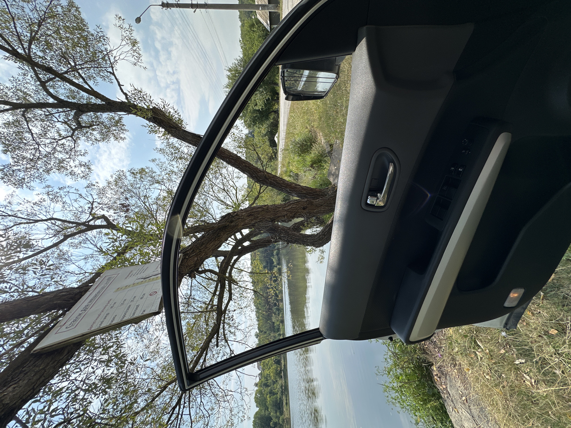 The image shows an open car door on the driver's side, parked on the grassy bank of a body of water, likely a river or lake, with trees and vegetation surrounding it. A white sign with text in a foreign language (likely Ukrainian) is attached to a tree, displaying a chart with measurements. In the background, a paved road runs parallel to the water, with more trees, houses, and a utility pole visible. The scene appears to be a scenic natural setting, possibly a recreational area, within the vicinity of Makiv, Ukraine. There are no people or animals visible in the image.