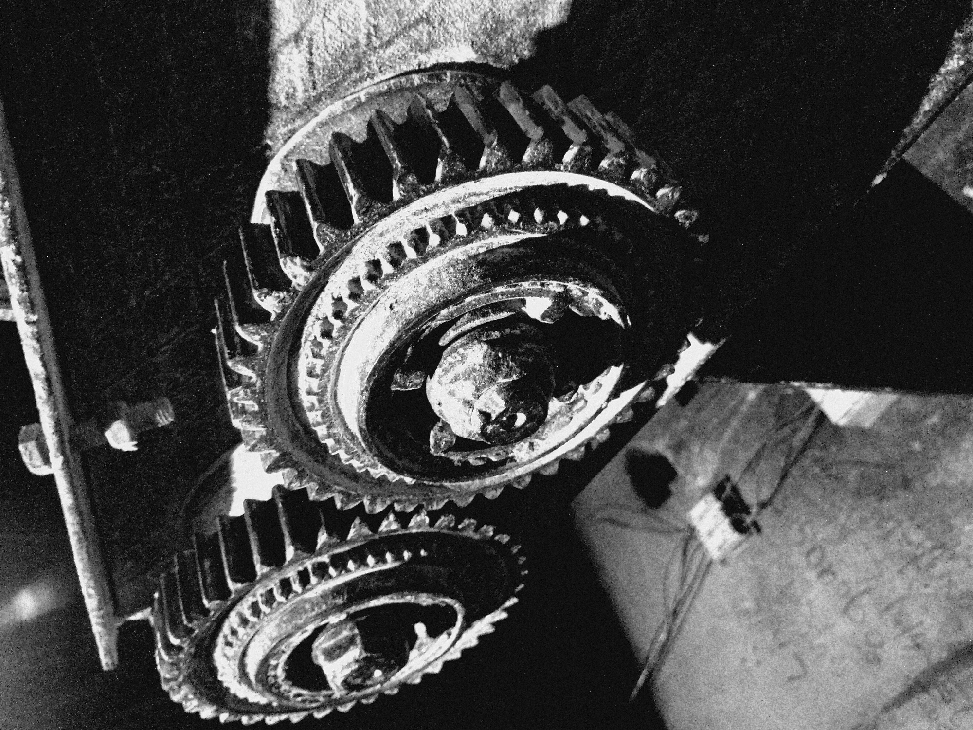 The media file presents a high-contrast, black and white close-up photograph of two large, heavily-toothed gears, likely part of an industrial mechanism. The image is captured from an oblique, slightly downward perspective, highlighting the robust and worn textures of the machinery.
The upper gear dominates the central-upper portion of the frame, showcasing its large, rugged teeth and an intricate inner ring featuring smaller, decorative or functional protrusions. A central bolt or shaft, showing signs of wear and corrosion, is visible at its core. Partially visible below and to the left is a second, similar gear, its structure mirroring that of the upper one, with visible teeth and an inner ring. Both gears appear to be made of heavy metal, possibly cast iron or steel, and display significant signs of age, wear, and surface texture, consistent with heavy use.
The lighting is dramatic and directional, creating stark contrasts between brightly illuminated metallic surfaces and deep, rich shadows. This strong illumination emphasizes the three-dimensional form and rugged details of the gears. On the left side, a metallic structural component, possibly a frame or housing, is visible, secured by a bolt. The background primarily consists of dark, abstract shadowed areas. However, in the lower right, a textured surface—likely concrete or another worn industrial material—is discernible. Faint, indistinct markings or text can be vaguely seen on this surface, but they are too blurry and incomplete to be clearly deciphered.
The image conveys a sense of powerful, industrial machinery, aged but enduring. There is no explicit activity depicted; the gears are static, captured in a moment of stillness, emphasizing their form and texture within their mechanical setting.