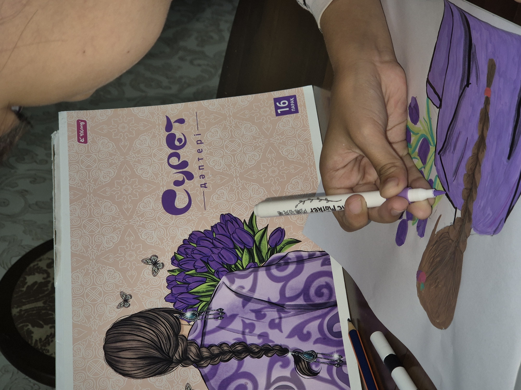 In an indoor setting, possibly a home or studio in Kokshetau, Kazakhstan, a person's hand is holding a purple marker and coloring a drawing. The drawing depicts a person with brown braided hair and a purple garment, with tulips in the background. The drawing is on a white sheet of paper, which is placed on a dark wooden table. In the background, there is a book or coloring book with a decorative pink cover featuring a woman with braided hair, tulips, and butterflies. The book's title appears to be in a foreign language. The overall scene suggests a creative activity, likely coloring or drawing, in progress. The lighting appears to be natural, coming from an unseen window.