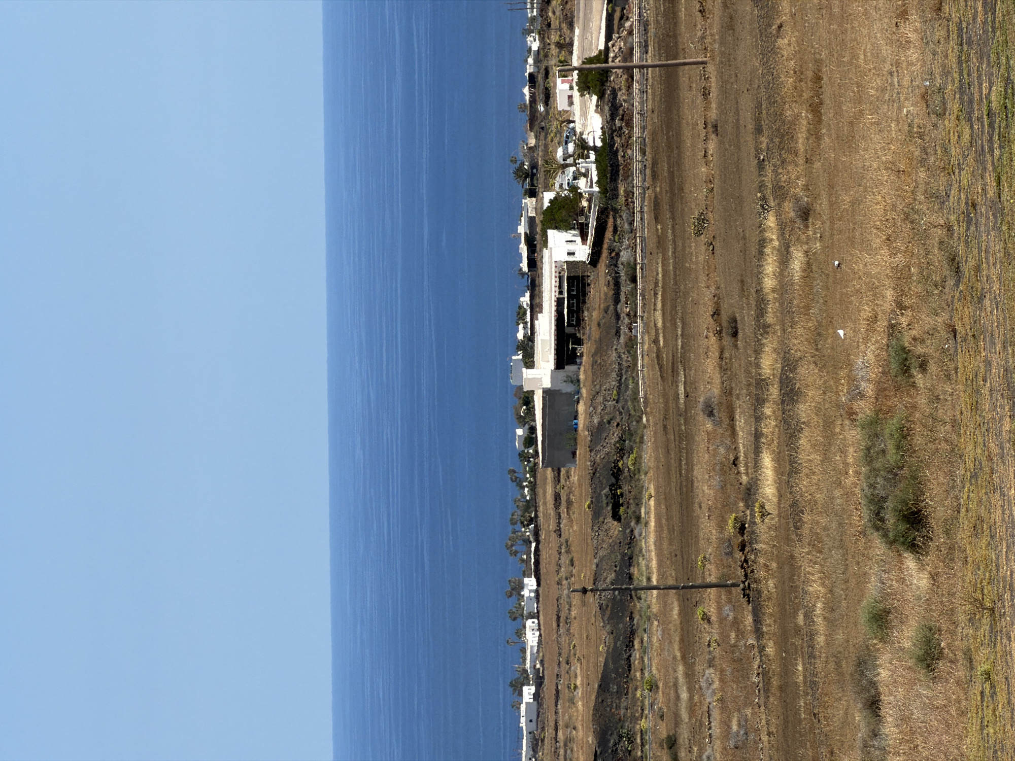 The image presents a panoramic coastal landscape, likely captured from an elevated perspective looking towards the sea. The left side of the frame is dominated by a vast, clear, light blue sky that meets a deep blue, calm sea at a distinct horizon line. Subtle ripples or current patterns are visible on the water's surface.
Along the coastline, a strip of land hosts several clusters of white buildings with flat roofs, characteristic of Mediterranean architecture. These structures, possibly residential or tourist accommodations, are interspersed with sparse green vegetation, including a few palm trees. A road or path appears to run parallel to the coast behind these buildings.
To the right of the coastline, a wide expanse of dry, arid terrain stretches into the foreground. This landscape is characterized by yellowish-brown earth, sparse, dry, straw-colored grass, and low-lying scrub, with some darker, rocky patches visible. In this dry field, a few dark, slender utility poles or fence posts stand vertically, and a wire fence runs horizontally along a section of the field. Two small white specks, possibly birds or debris, are discernible on the dry ground in the midground.
The scene depicts a bright and clear sunny day, with strong, even lighting suggesting midday. No specific human or animal activity is visible. The combination of white architecture, a deep blue sea, and an arid volcanic landscape is highly consistent with the location of Tías, Spain, particularly on an island like Lanzarote. There is no visible text in the image.