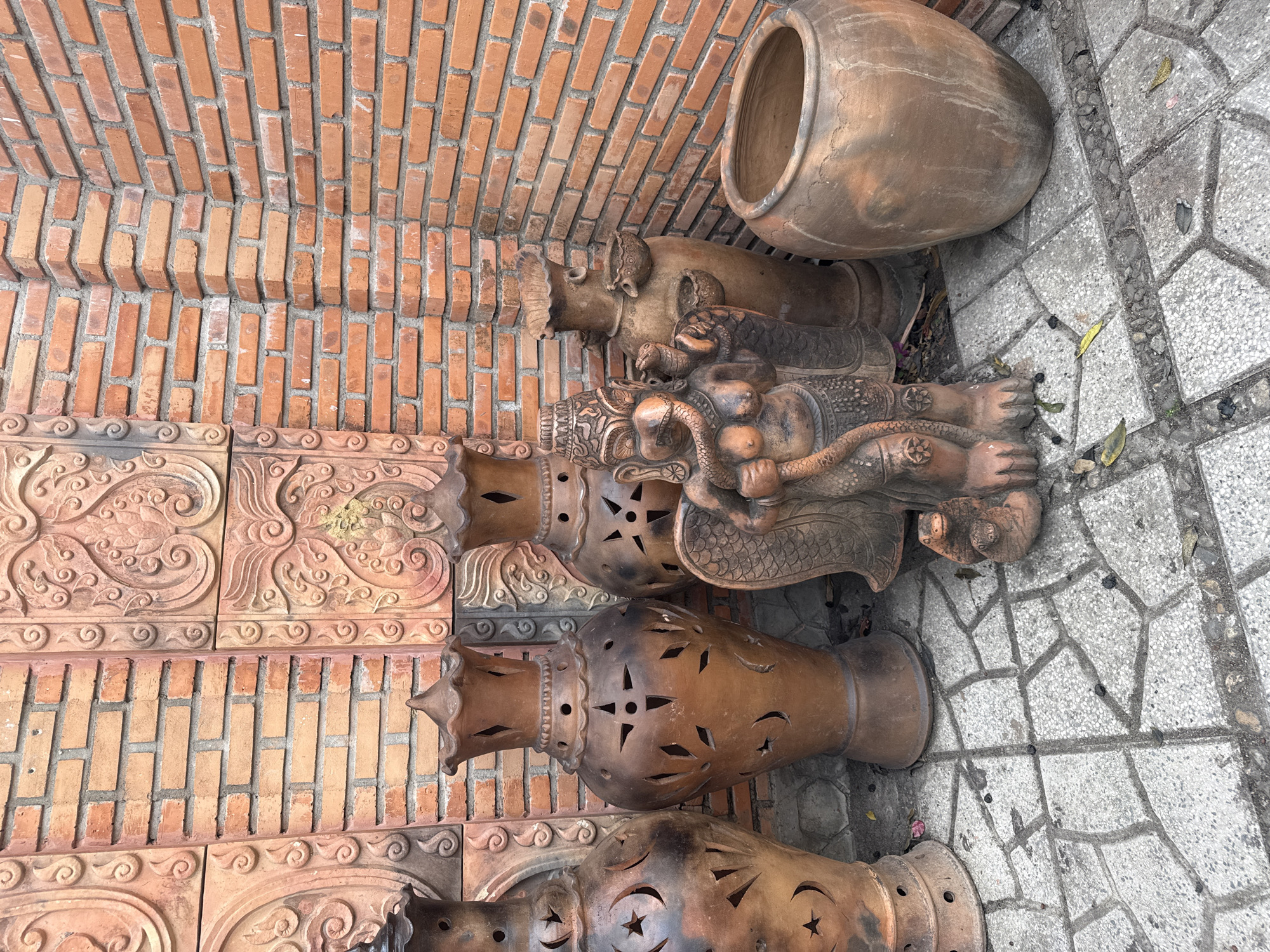 In Bac Nha Trang Ward, Vietnam, several terracotta objects rest on a weathered, stone-tiled ground adjacent to a brick wall. A large, round, earthenware jar sits near an arrangement of decorative clay pottery. These include intricately carved tiles and hollowed, conical-shaped objects with star-shaped perforations. Atop these is an elephant figurine and a reclining human-like figure entwined with a serpentine element. The overall arrangement displays a collection of artistic and functional clay items.