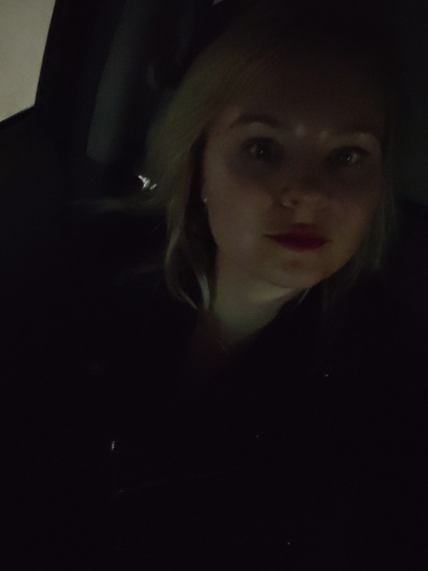 A dimly lit, close-up shot features a woman, possibly taking a selfie, inside what appears to be a car at night. The woman, with light blonde hair, is looking directly at the camera with wide, open eyes. She is wearing bright red lipstick and has a subtle nose piercing. A faint necklace is visible around her neck. Her face is partially illuminated, creating highlights on her forehead and nose, while the rest of her body and the background are cast in deep shadow. She is wearing a dark top or jacket. The overall setting suggests a private moment within an enclosed space during nighttime hours, with artificial light source illuminating her face. No specific details about the location are visible in the image, but the provided context indicates the location is Izhevsk, Russia. There is no visible text.