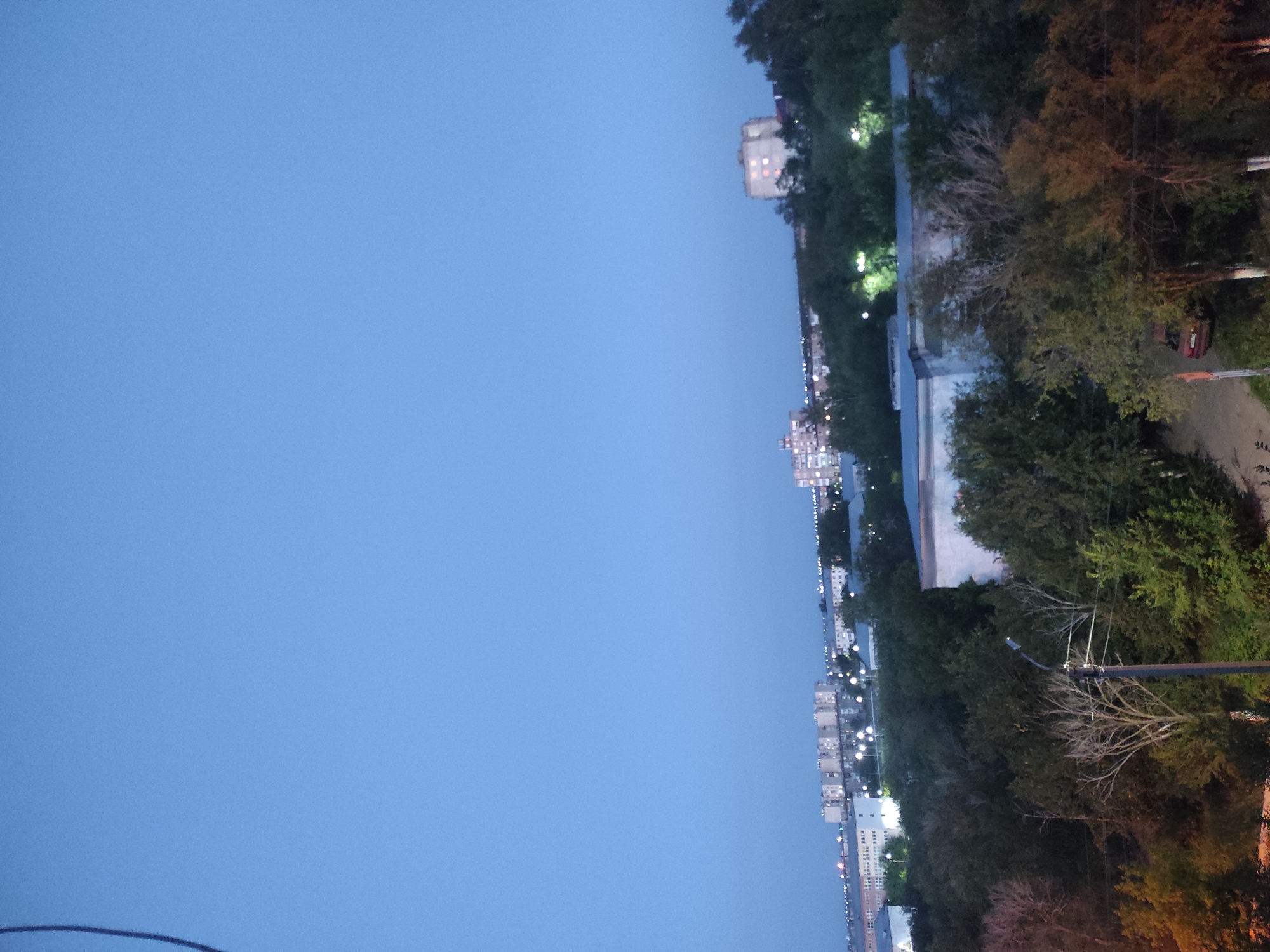 The image depicts a twilight view of a city, likely Shakhtinsk, Kazakhstan, characterized by a vast expanse of clear, pale blue sky dominating the upper portion. Below the sky, a low-rise urban landscape unfolds, featuring several multi-story apartment buildings and other structures. Streetlights are visible, casting a warm glow across the scene, indicating the transition from day to night. Dense foliage, composed of trees and bushes, partially obscures the buildings, creating a sense of depth and natural integration within the urban environment. A single red car is visible on a dirt road in the foreground, suggesting residential or less-trafficked areas. The overall impression is one of a quiet, developing city settling into the evening.