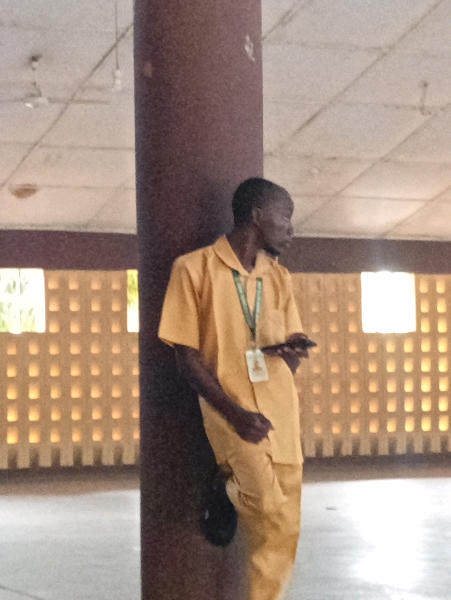 A dark-skinned young man wearing a yellow uniform shirt and pants leans against a thick, dark brown pillar. He is looking to his left and holding a dark-colored smartphone in his right hand. A white identification card with a picture is attached to a green lanyard around his neck. The background features a cream-colored ceiling with exposed lighting fixtures and what appears to be a ventilation system. Behind the man, a wall is visible with a repeating pattern of illuminated rectangular openings. The overall setting suggests an indoor public space, possibly a school or administrative building, likely located in Gombe, Nigeria, given the provided context.