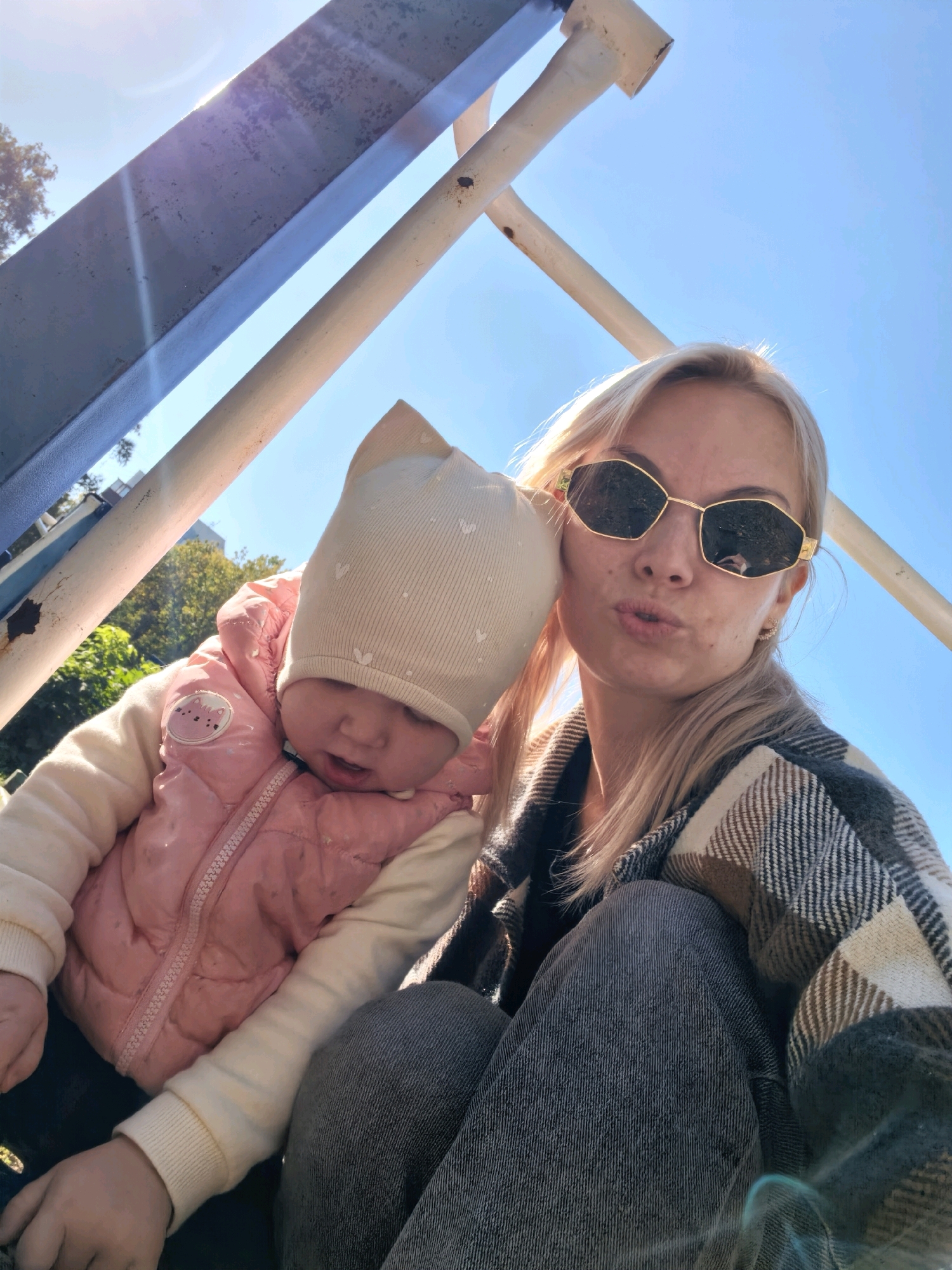 A woman and a baby are seated outdoors on a sunny day, seemingly on a playground structure. The woman, with blonde hair and sunglasses, is in the foreground, leaning her head towards the baby. She is wearing a plaid jacket. The baby, wearing a hat and a pink puffer vest over a cream-colored long-sleeved shirt, is positioned to the woman's left, looking downwards. The background shows a clear blue sky and some greenery, with parts of a metal structure visible, suggesting a playground or outdoor recreational area. There is no specific indication of the city of Sterlitamak, Russia within the image itself.