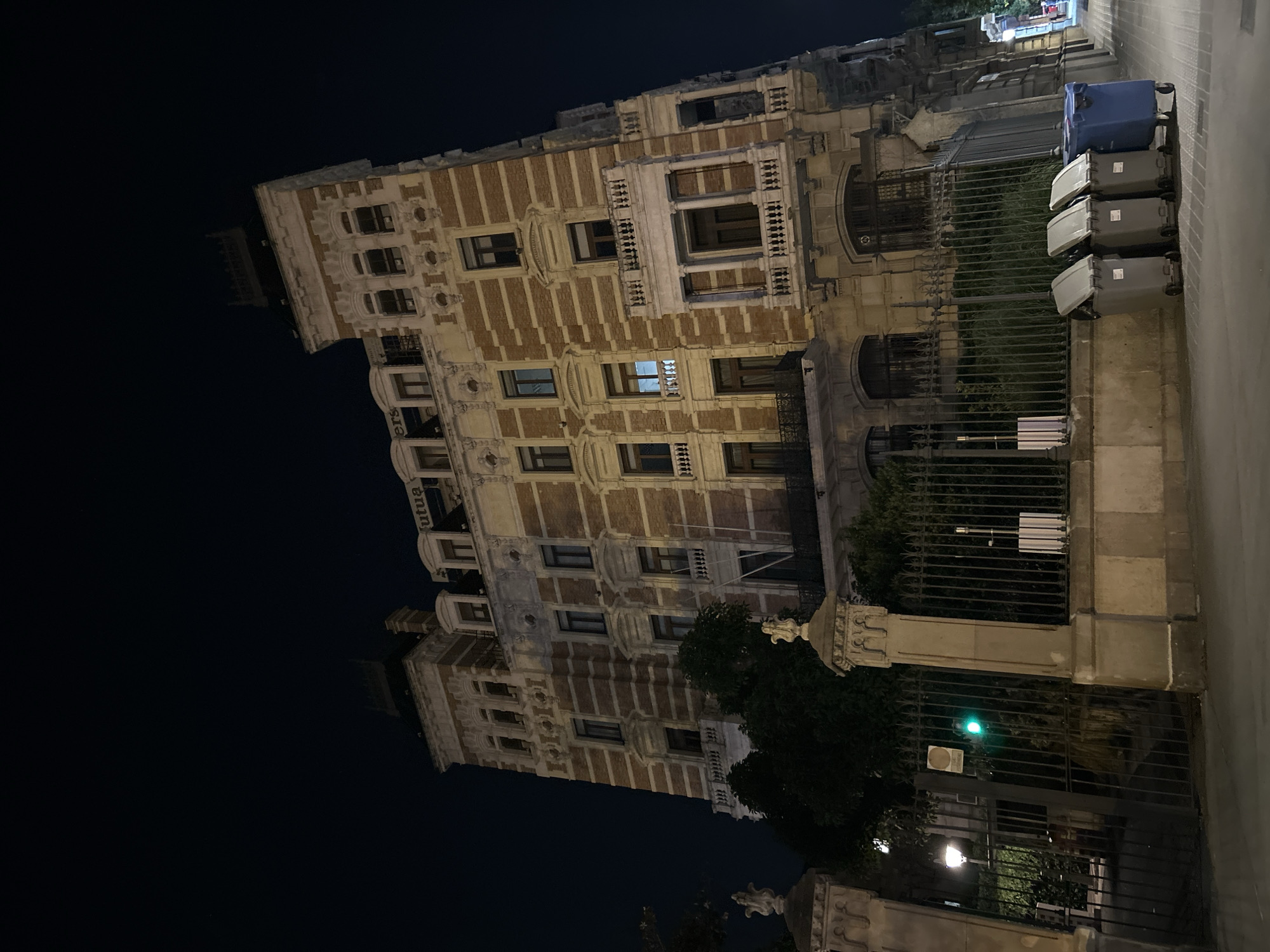 At night, a tall, ornate building stands next to a sidewalk in Barcelona, Spain. The building, primarily brick with white architectural details, has multiple floors with windows. A single light glows from one of the windows. A black metal fence runs along the building's side, partially obscured by trees and bushes. A green light is affixed to the fence. To the right of the fence, trash bins are stacked on the sidewalk. The sky in the background is dark.