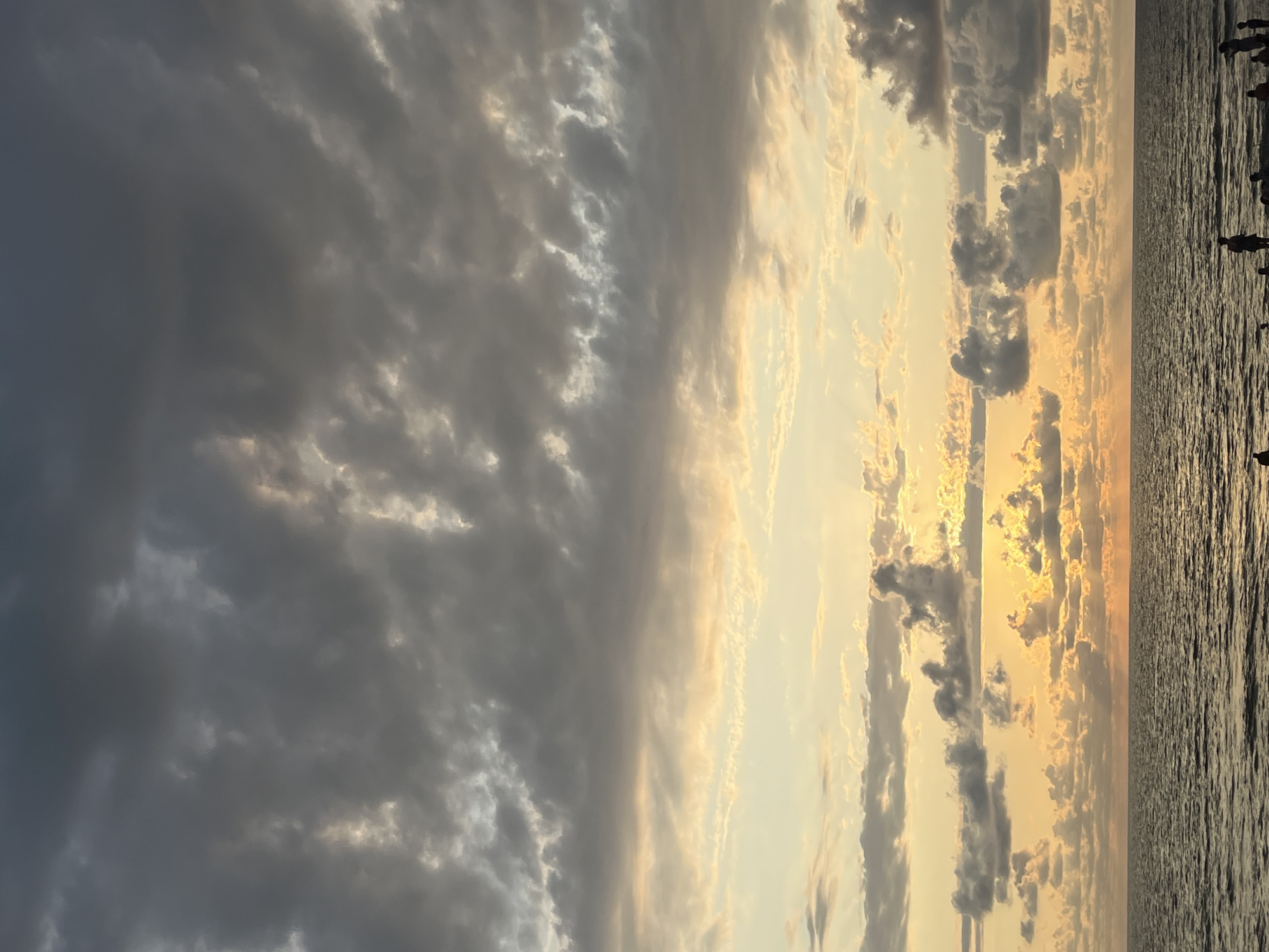 This image depicts a serene sunset scene at Ureki Beach, Georgia, characterized by the Black Sea meeting a sky filled with dramatic clouds. The sky transitions from a soft yellow and orange near the horizon to darker, brooding gray clouds higher up. Sunbeams pierce through the clouds, creating a textured effect. The ocean is calm, with gentle waves reflecting the colors of the sunset. Along the shoreline, several silhouetted figures can be seen, suggesting a small gathering of people enjoying the view. The overall atmosphere is tranquil and picturesque.