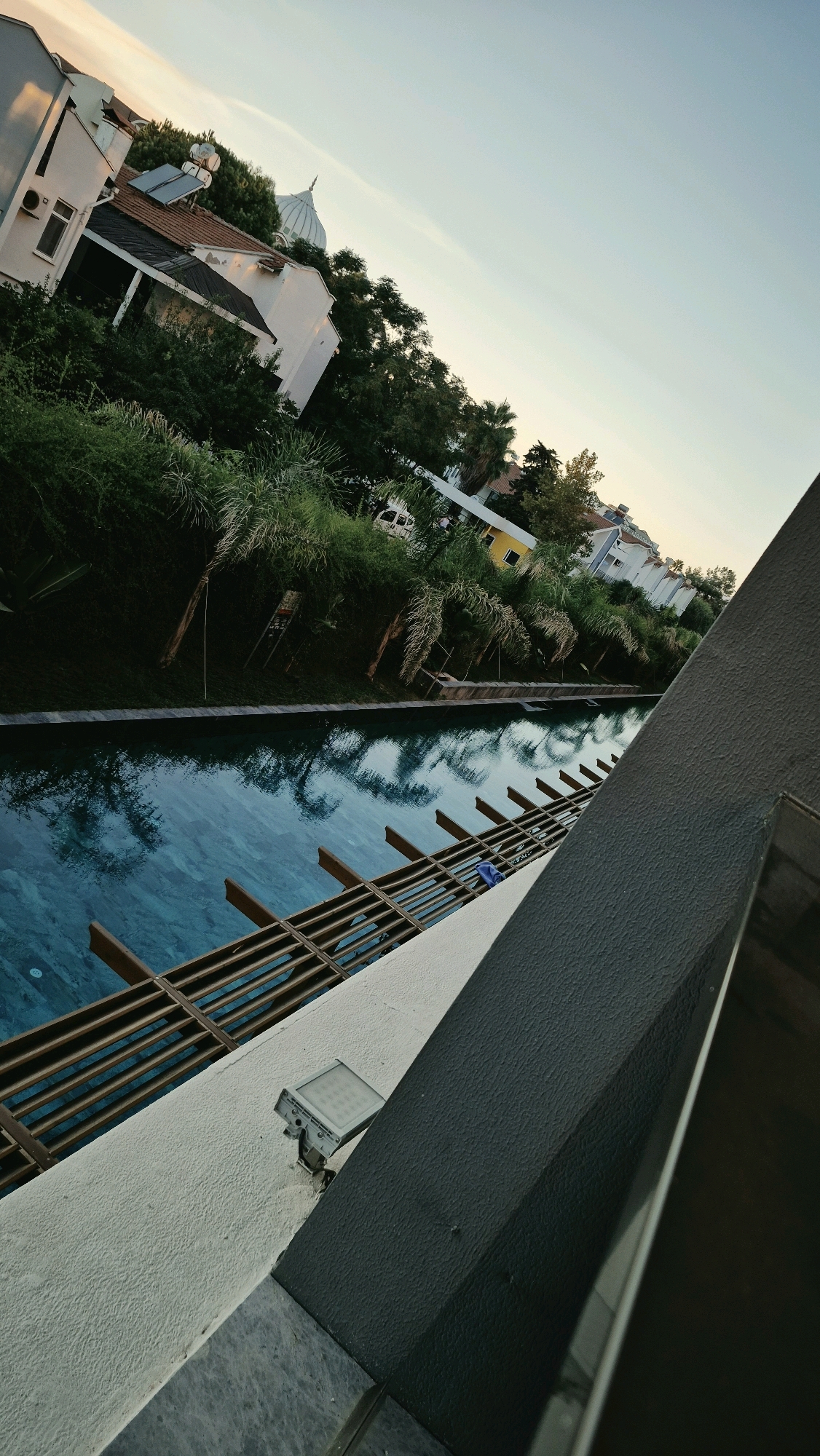 An angled, medium shot captures a swimming pool area in Serik, Turkey, during the late afternoon. The water, a vibrant blue, is calm and reflects the surrounding palm trees and buildings. A wooden deck structure runs alongside the pool, partially submerged in the water. In the background, white and yellow buildings are visible, nestled amongst lush green trees and palm trees. A prominent white dome, possibly part of a religious building, stands out against the clear, pale blue sky. A security light is mounted on a textured white wall in the foreground. The image has a slightly tilted perspective, adding a dynamic feel to the scene.