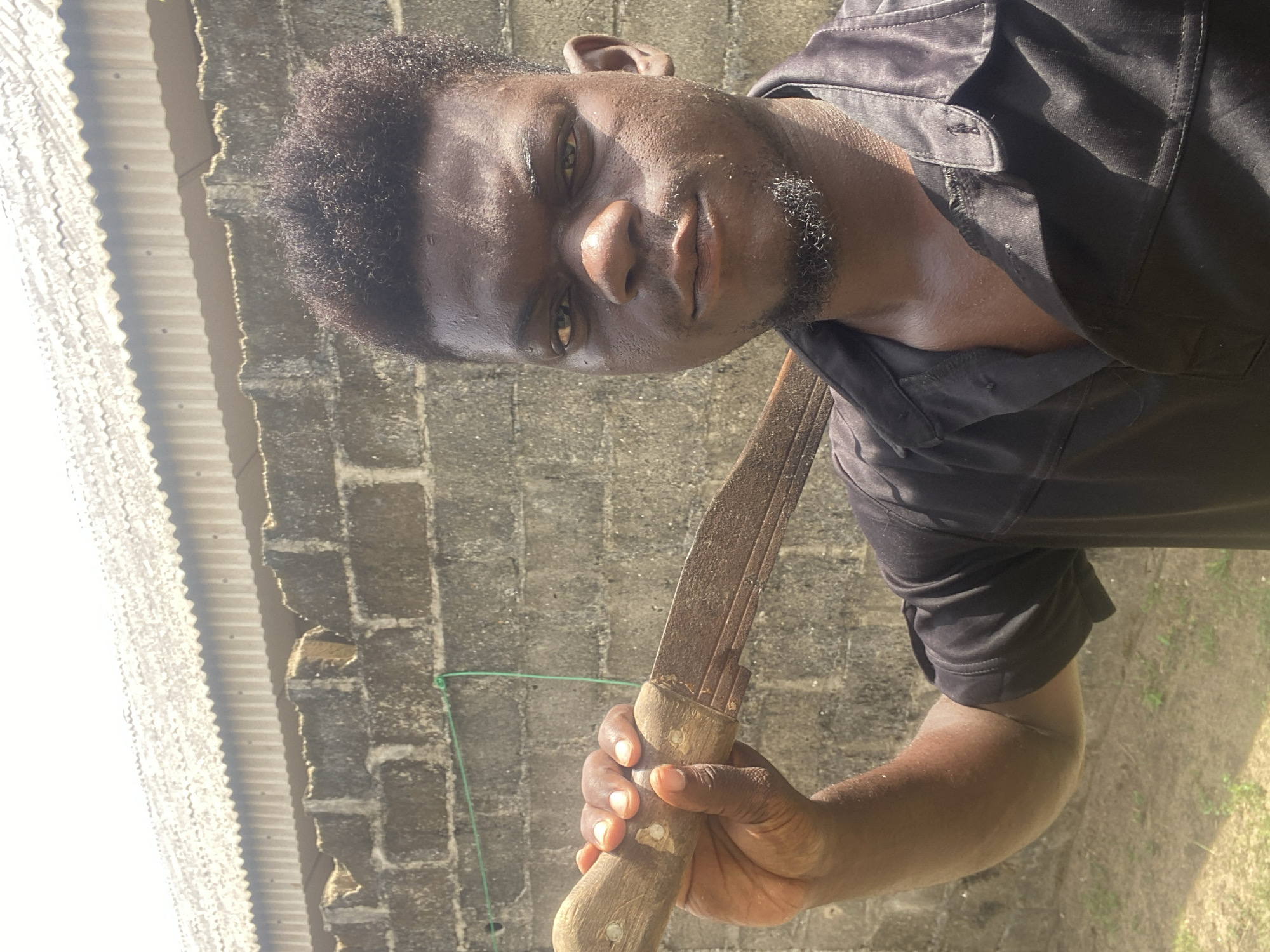 A person stands in front of a building with a corrugated metal roof and a block wall. The person is holding a large knife or machete with a wooden handle. The location appears to be outdoors in a city marked Unknown, Unknown. The individual is facing the camera. The image's perspective is slightly tilted.