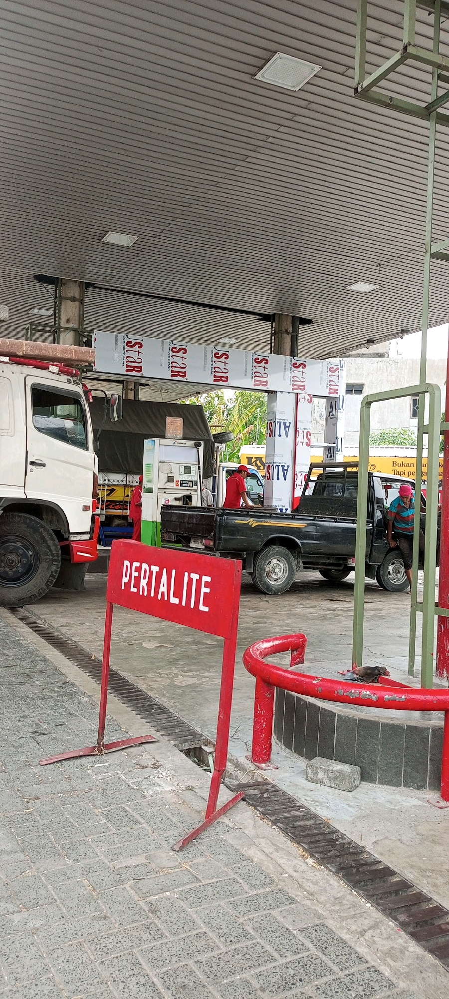 In Plaosan, Indonesia, a gas station scene unfolds under a corrugated metal roof supported by pillars. A white truck is parked on the left, its large wheel visible. To its right, a black pickup truck is positioned, with a man in a red shirt visible in its bed, seemingly attending to something. Near the pickup truck, another individual wearing a striped shirt and pink head covering stands by the open passenger door.
In the foreground, a red sign with white lettering reads "PERTALITE," a type of fuel. This sign is supported by two red metal stands. The ground is paved with interlocking grey bricks, with a metal drainage grate running along a section of it. Behind the vehicles, a white gas pump station with digital displays is visible, with a person in red clothing standing behind it. Decorative panels with "J-STAR" and "ALUS" branding adorn the pillars supporting the roof. The environment appears to be outdoors with some greenery visible in the background.