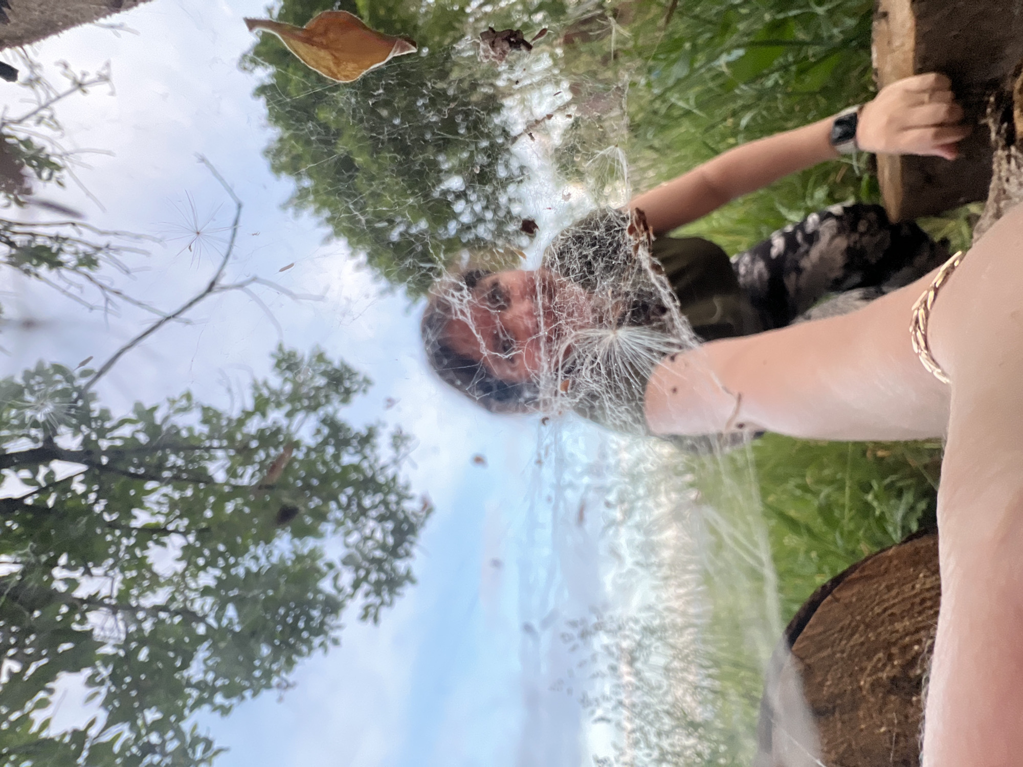 The image depicts a person outdoors, with a focus on a spiderweb in the foreground. The spiderweb is large and intricate, with debris and some plant matter caught in it. Through the web, a person is visible in the background, appearing to be in a seated or crouched position amidst green foliage. The person is wearing a dark-colored shirt and patterned pants. There are logs or pieces of wood visible in the lower part of the frame, suggesting an outdoor, possibly wooded or garden setting. The sky above is partly cloudy. The image appears to be taken from a low angle, looking up towards the person and the trees. No specific landmarks or identifiable features of Panteleevka, Russia are discernable.
