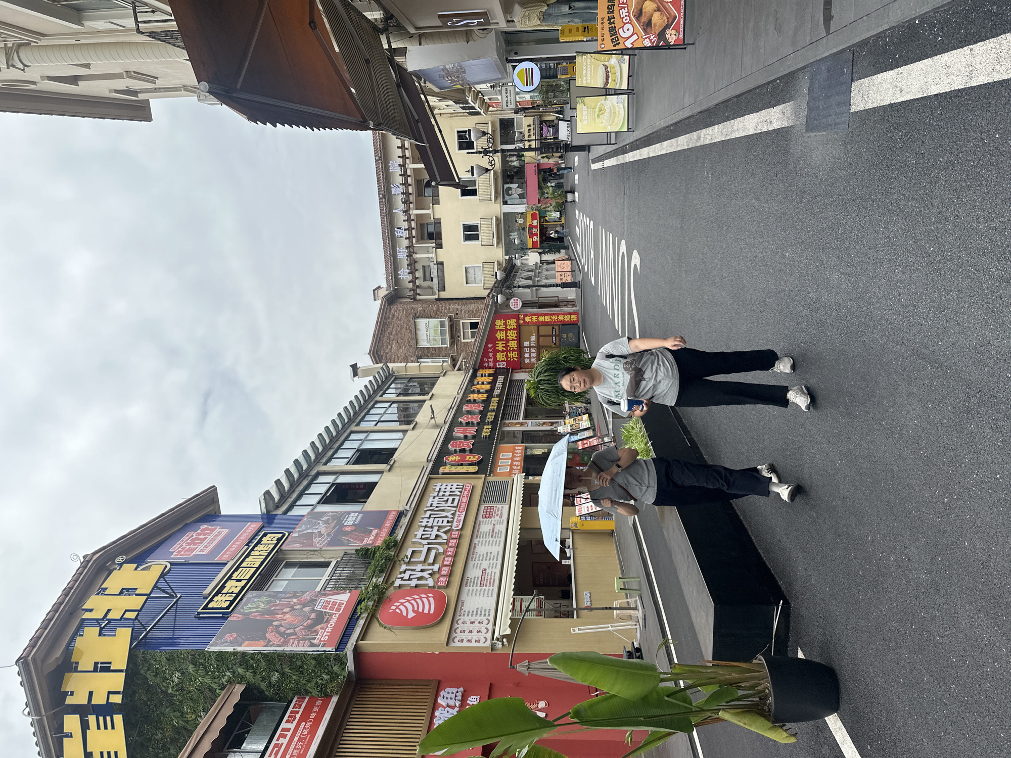 Here's a detailed description of the image:
**Overall Scene:** The image shows a street scene with several buildings lining the side. Two people are walking on the street. The setting appears to be an urban area, likely a commercial district, with a variety of shops and restaurants.
**Location:** The image is located in the city of Chongqing High-tech Zone, China. This is based on the presence of Chinese characters on the storefront signs.
**People:** Two people are in the image. One person is holding an umbrella.
**Objects:**
* Buildings: Varying heights with shopfronts displaying signage in Chinese.
* Signage: Numerous shop signs with a mix of Chinese and potentially English text.
* Umbrella: Held by one of the people.
* Plants: A potted plant.
* Road markings: A white line.
**Actions/Interactions:**
* People walking along the street.
**Environment:**
* Weather: Overcast sky.
* Road: Asphalt street.
* Urban setting.