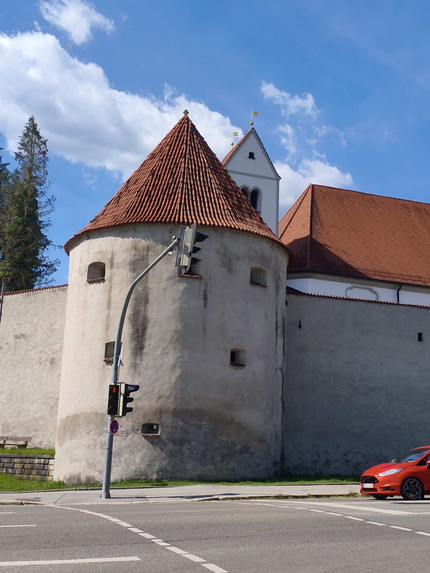 This is an eye-level, medium shot of the St. Mang's Abbey in Füssen, Germany, on a clear, sunny day. The abbey complex features a round tower with a conical red-tiled roof and a white church spire with two small flags. A grey wall surrounds the buildings. In the foreground, an orange Ford Fiesta is parked at a traffic light, which is red. A traffic sign indicating "no stopping" is visible below the traffic light. The street in front of the abbey has white lane markings and a pedestrian crossing. Green grass and a stone bench are visible on the left side of the image.