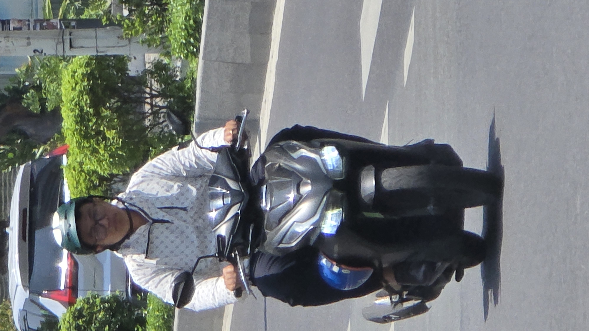 A medium shot captures a person riding a scooter on a street with a car parked along the edge, likely in Nha Trang, Vietnam. The person, possibly male, wears glasses and a helmet and is clad in a light-colored, patterned long-sleeved shirt. The scooter is dark and features reflective silver accents. A parked silver-colored car is located on the left side of the frame, near lush green plants and shrubbery, and an unidentified sign that is attached to a tall pole behind the green plants. The street is made of light-colored asphalt. The scene is bathed in bright sunlight, evidenced by sharp shadows cast by the scooter and its rider.