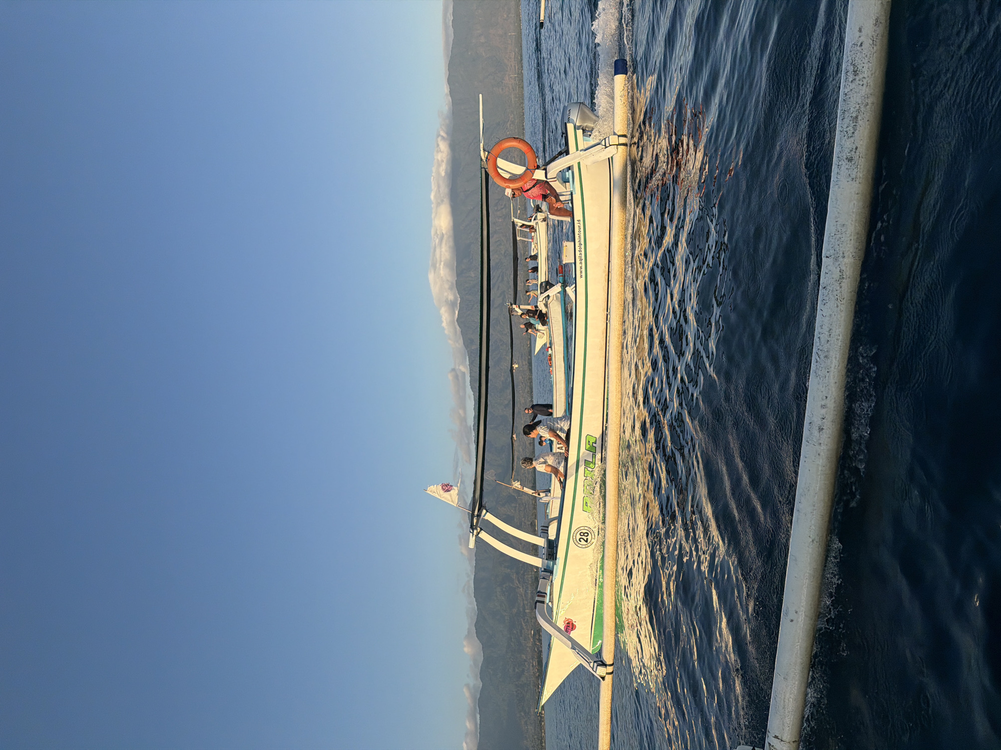 In an Indonesian coastal landscape, a narrow white boat with green accents is cutting through the dark blue water, leaving a foamy wake. The boat, identified with the number 28 and the word "PRILA" in green lettering, is carrying several passengers. A woman in a white top and dark shorts sits on the left side of the boat, looking forward. Next to her, another woman in a white patterned top and dark shorts also looks ahead. Further back, a person wearing a pink shirt and a hat is seated on the right side, looking towards the viewer. A person in a black shirt and pants stands at the stern of the boat. The boat is equipped with an orange life preserver hanging from a support structure. A white flag with a purple emblem flutters from a pole at the front of the canopy. In the distance, a mountainous coastline is visible under a clear blue sky with a few scattered clouds. Another similar boat is partially visible to the left. The water is rippling, reflecting the golden hues of the setting sun.