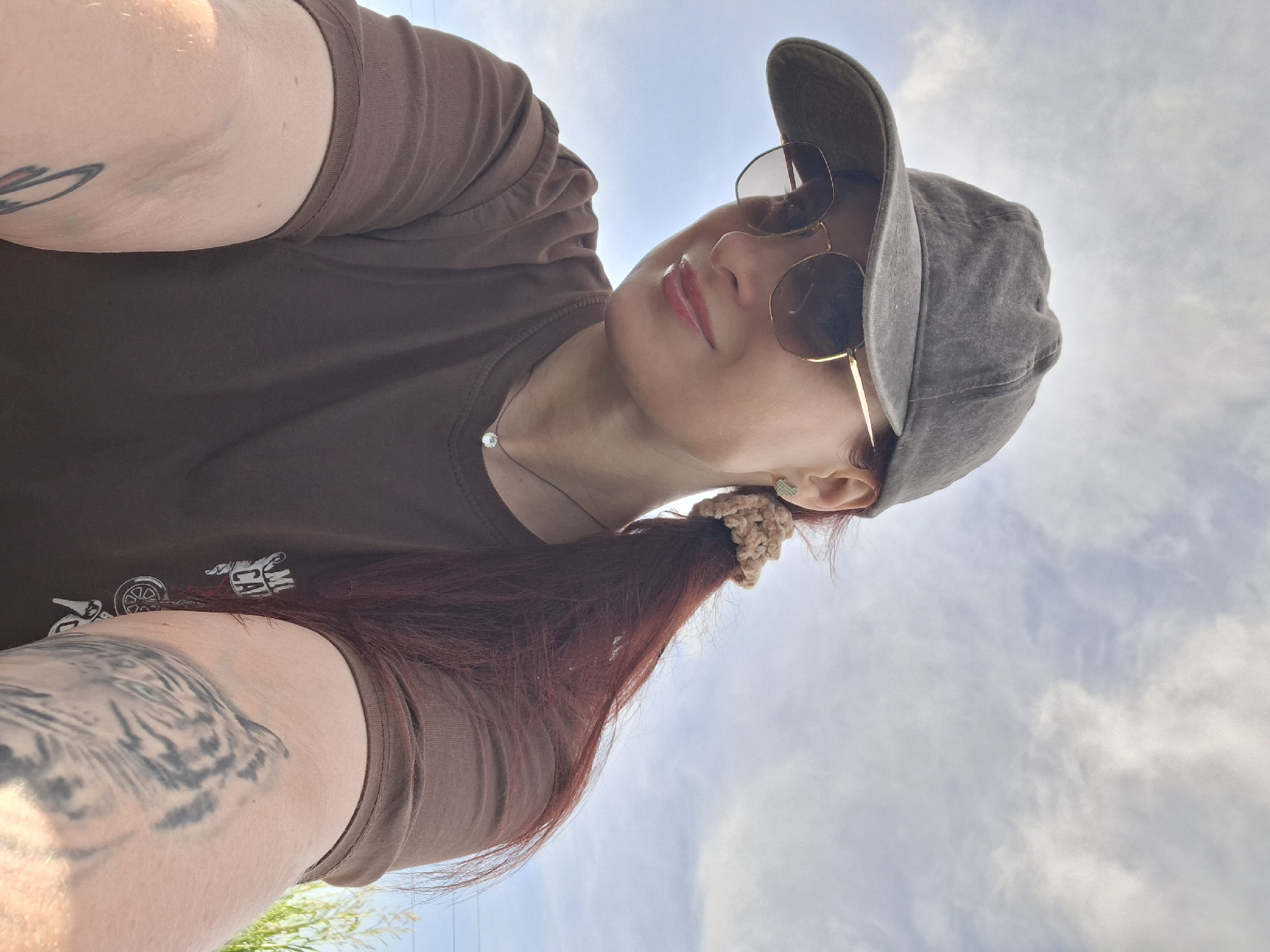 A low-angle selfie captures a person, likely female, looking upwards against a bright, partly cloudy sky. The subject's head is tilted to the right, and their body fills the left side of the frame.
The person is wearing a plain brown t-shirt. On their right arm (viewer's left), a detailed tattoo of a tiger or large cat's face with light green eyes is prominently visible. A portion of another tattoo is visible on their left arm. The t-shirt also features a faint graphic on the chest with what appears to be motorcycle-related imagery and some text, including the letters "MA" and "CAM."
They are wearing a faded grey baseball cap and large, round sunglasses with gold frames that cover their eyes. Their lips are slightly parted and appear to have a glossy finish. A simple necklace with a single clear bead or gem is visible around their neck. Long, reddish-brown hair is pulled back into a ponytail, secured with a textured light brown or beige scrunchie. A small, sparkling earring is visible on their right ear.
The background is a clear, bright sky with soft, scattered white clouds, indicating sunny outdoor conditions. A sliver of green foliage and faint utility lines are visible at the very bottom edge of the image. The overall mood is casual and relaxed.
The location context is Забалоцкі сельскі Савет, Belarus.