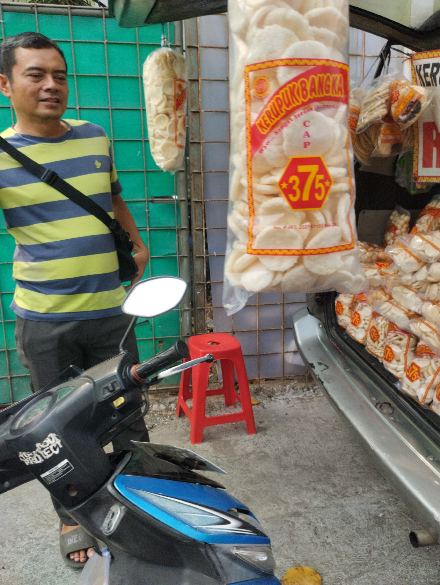 A man stands next to a parked motorcycle in front of a stall selling packaged crackers in Bekasi, Indonesia. The man, wearing a blue and yellow striped t-shirt and grey pants, has a black bag slung across his chest. He is looking towards the left of the frame. The stall displays numerous clear plastic bags of crackers, many with red and yellow labels featuring the text "CAP 375". A bright red plastic stool is visible near the stall. The background consists of a green metal grid fence. The motorcycle is black and blue, with a silver mirror and handlebars. The ground is concrete.