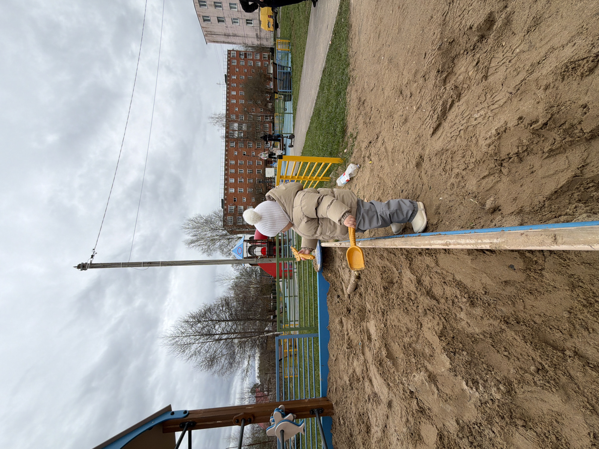 A child is playing in a sandbox in a playground, likely in Reshetnikovo, Russia, given the presence of typical Russian urban architecture in the background. The child, wearing a winter hat, jacket, and trousers, is holding a yellow plastic shovel. The sandbox has a blue and wooden border. A building with multiple windows is visible in the background, along with other playground equipment, including a climbing structure. The sky is overcast.