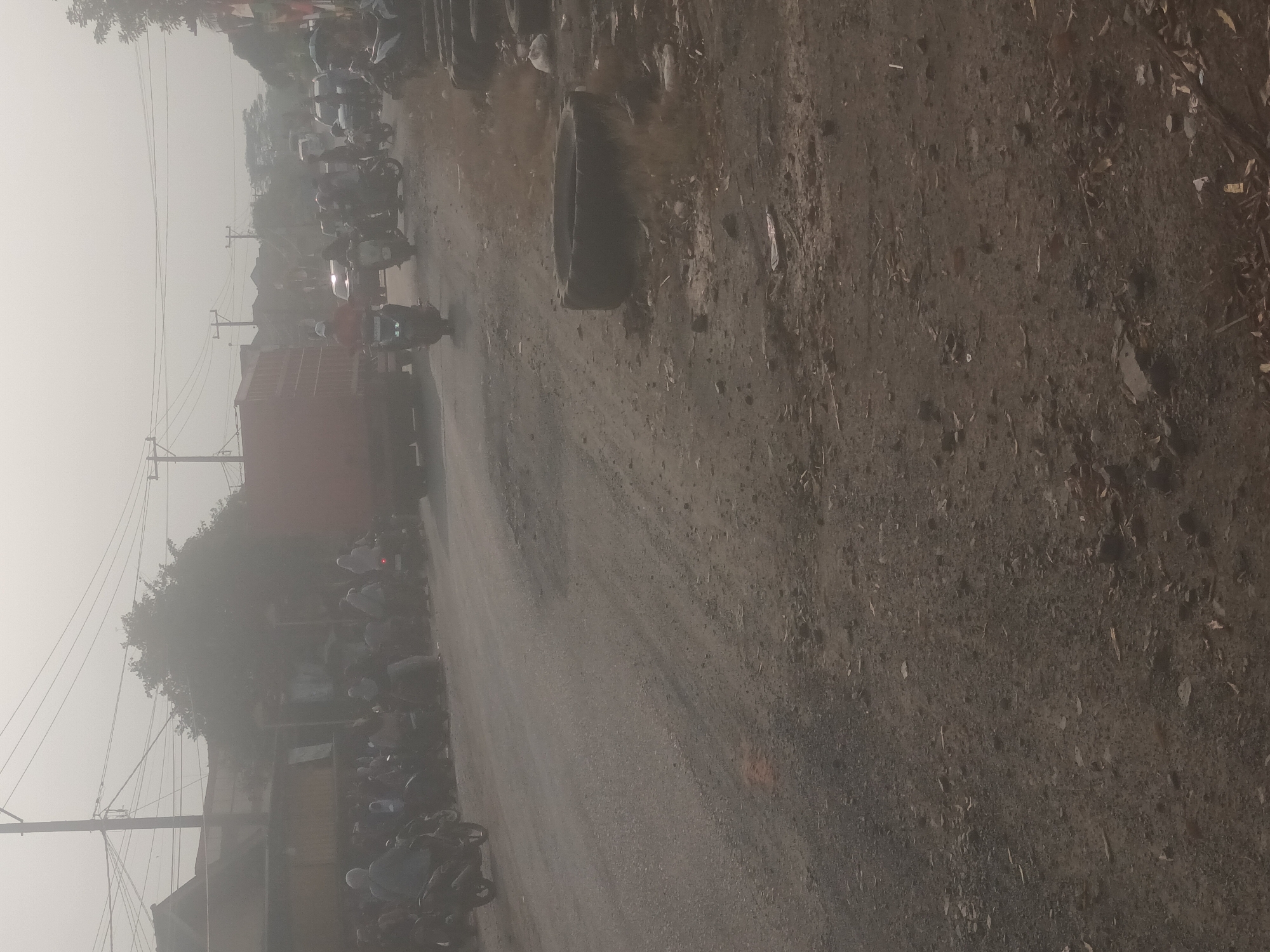 The image depicts a hazy street scene in Mulyo Asri, Indonesia, likely during the day given the lighting. A dusty, unpaved road dominates the foreground, littered with small stones and debris. To the left, a line of motorcycles is parked, with several individuals standing around them. Beyond the motorcycles, a large, red truck is visible, parked across the road and partially obscuring buildings in the background. Further into the background, more vehicles, including motorcycles and cars, are present on the road, suggesting traffic congestion. A few trees and utility poles with overhead wires are scattered throughout the scene. On the right side of the road, a discarded tire is prominently placed, and a small pile of trash is visible nearby. The overall atmosphere appears to be dusty and overcast, possibly due to weather conditions or pollution.