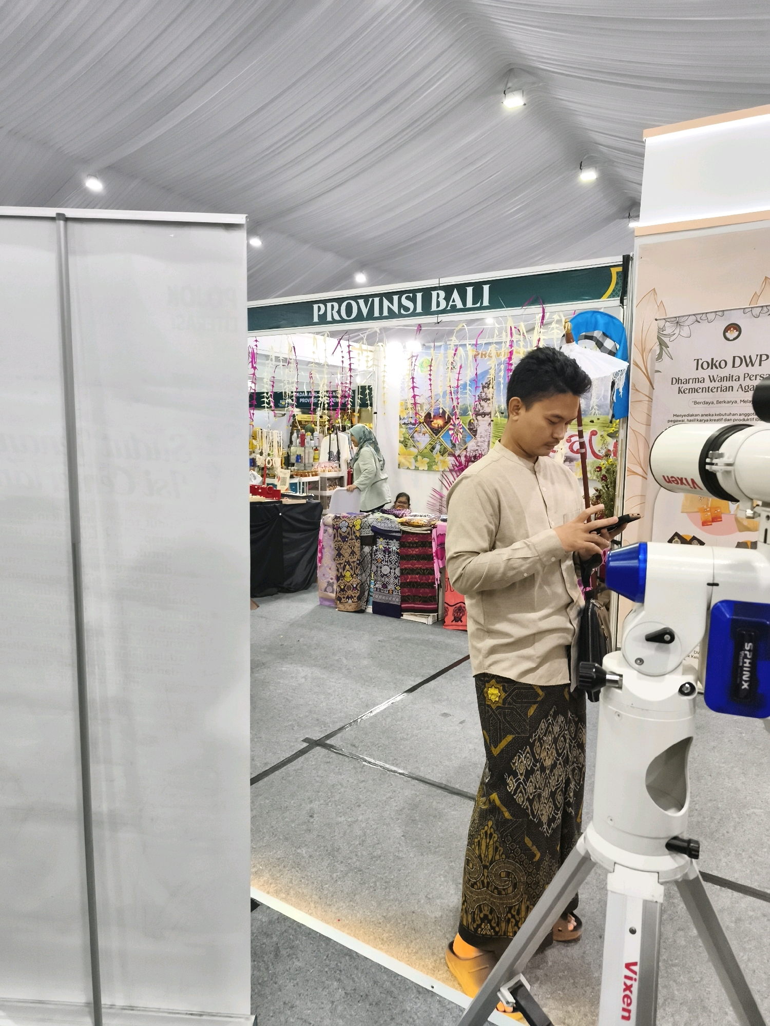 A man in a beige shirt and batik sarong stands indoors, looking at his phone. He is positioned in front of a white tripod-mounted telescope with a blue and white mount. Behind him, a banner reads "PROVINSI BALI". To his right, a display shows information for "Toko DWP Dharma Wanita Persatuan Kementerian Agama". In the background, a woman in a headscarf stands behind a stall with various items displayed. The floor is gray concrete, and the ceiling is draped with white fabric. The overall setting appears to be an exhibition or trade show. This event is being held in the city of Semarang, Indonesia.