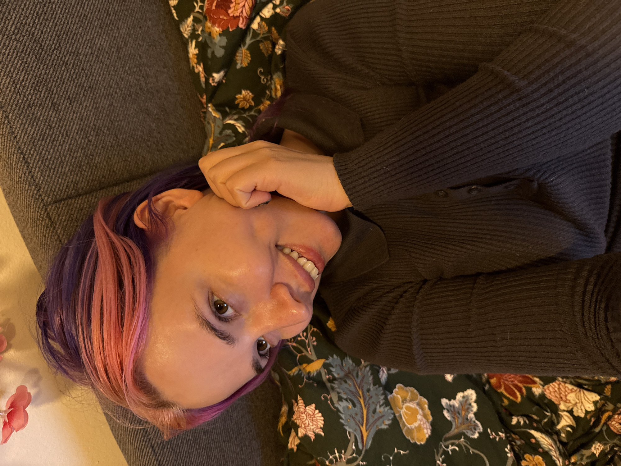 A woman lies on her back on a couch, turned 90 degrees counterclockwise. She has purple and pink hair, and is wearing a ribbed, long-sleeved dark gray sweater. She is smiling slightly, with one hand near her face. The couch has a dark gray fabric upholstery. Under her body there is a patterned fabric of dark green and floral elements. The location is not discernible.