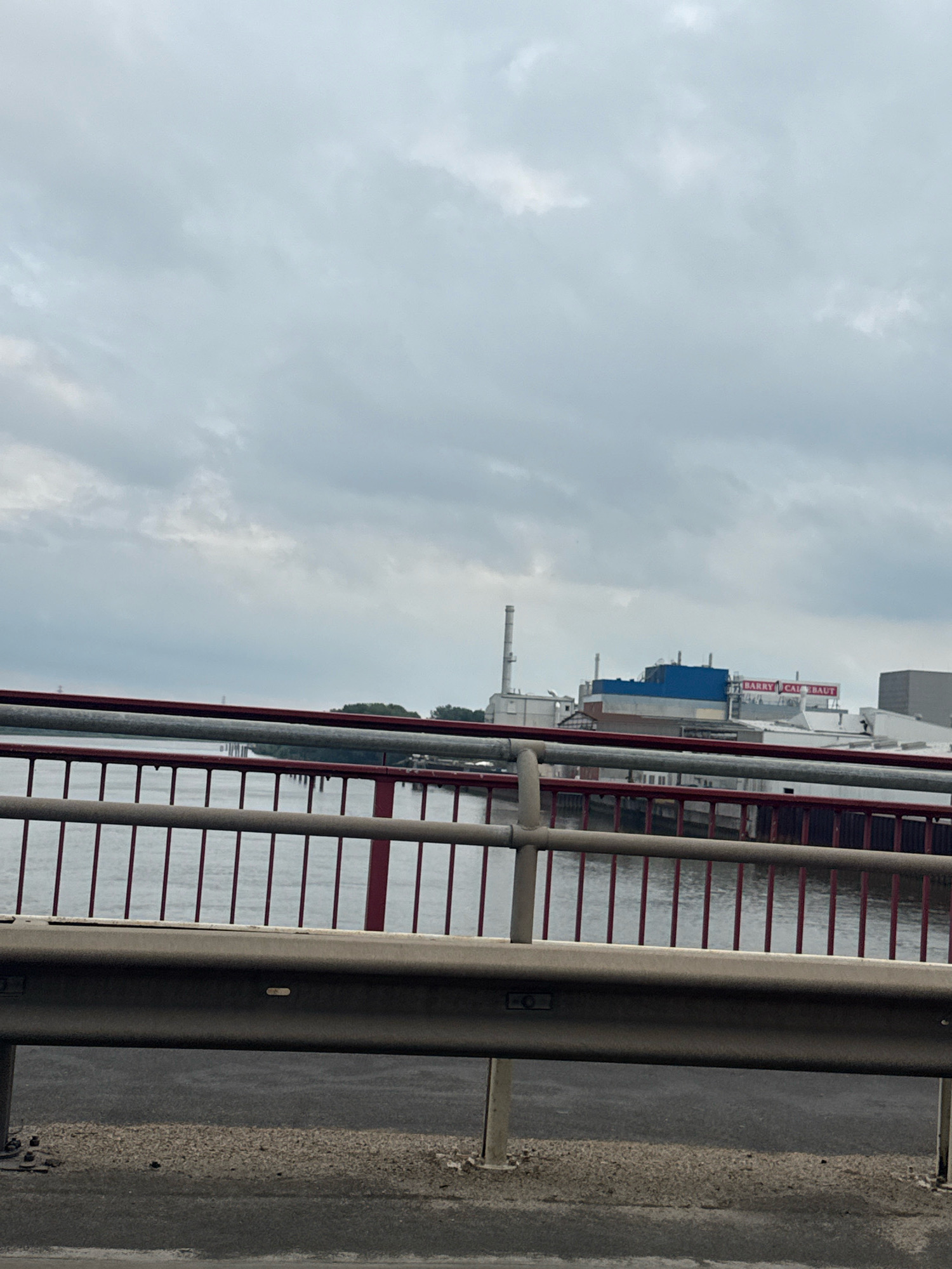 The image shows a cloudy sky over a body of water, likely a river or harbor, with industrial buildings in the background. In the foreground, a bridge railing is visible. The industrial buildings appear to be part of a factory or processing plant, with smokestacks and large structures. One building has a prominent blue section, and text on the facade reads "BARRY CALLEBAUT." This suggests the location is a Barry Callebaut chocolate factory, which has a facility in Hamburg, Germany, specifically in the Harburg district, known for its industrial presence. The water appears calm, and there are no people or vehicles clearly visible in the frame. The overall impression is a somewhat overcast, industrial waterfront scene.