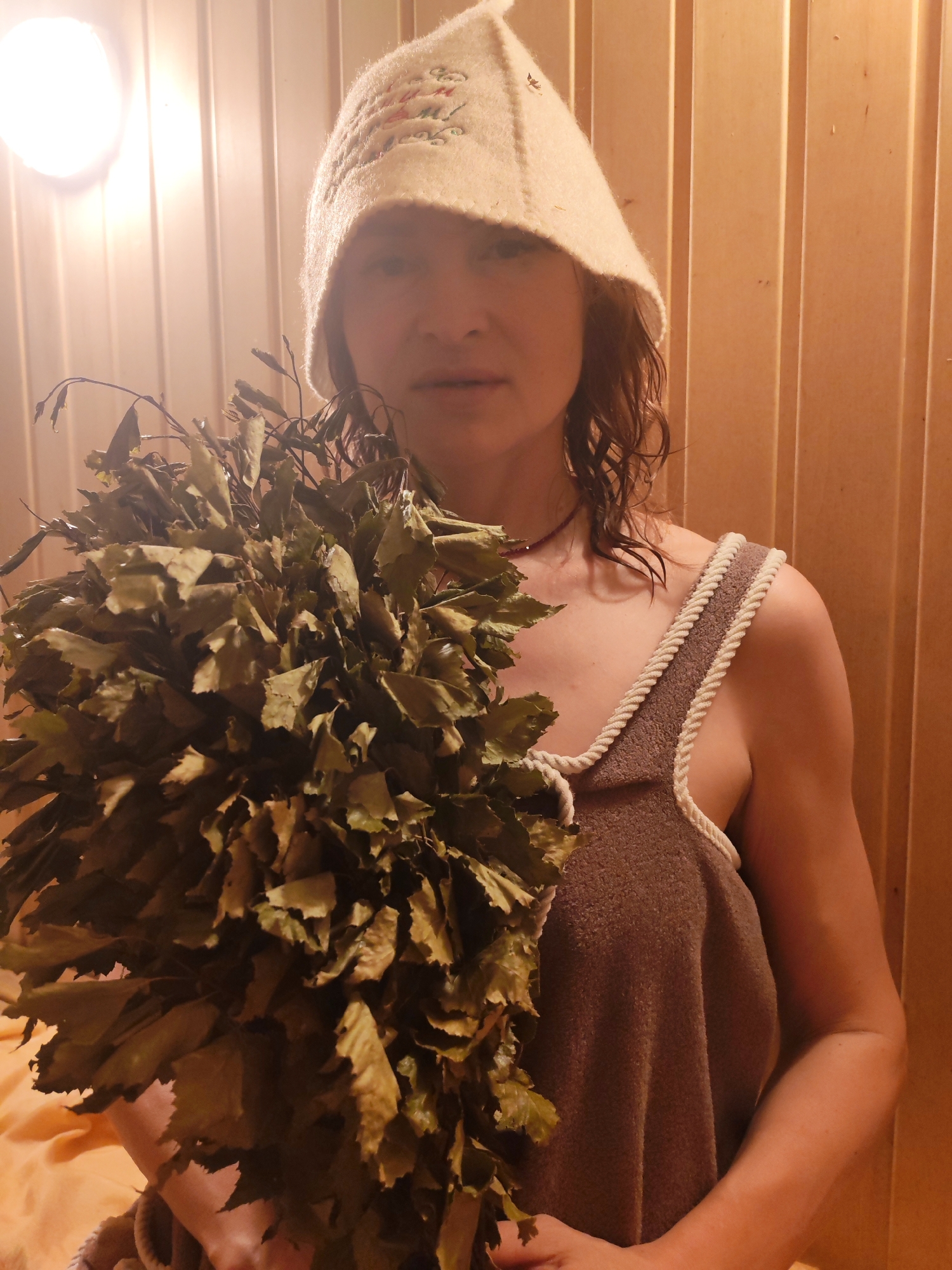 A woman is in a sauna. She is wearing a felt sauna hat and a brown towel dress. She is holding a bunch of leafy branches in front of her. The sauna is made of light brown wood. A light fixture is on the upper left side of the image.