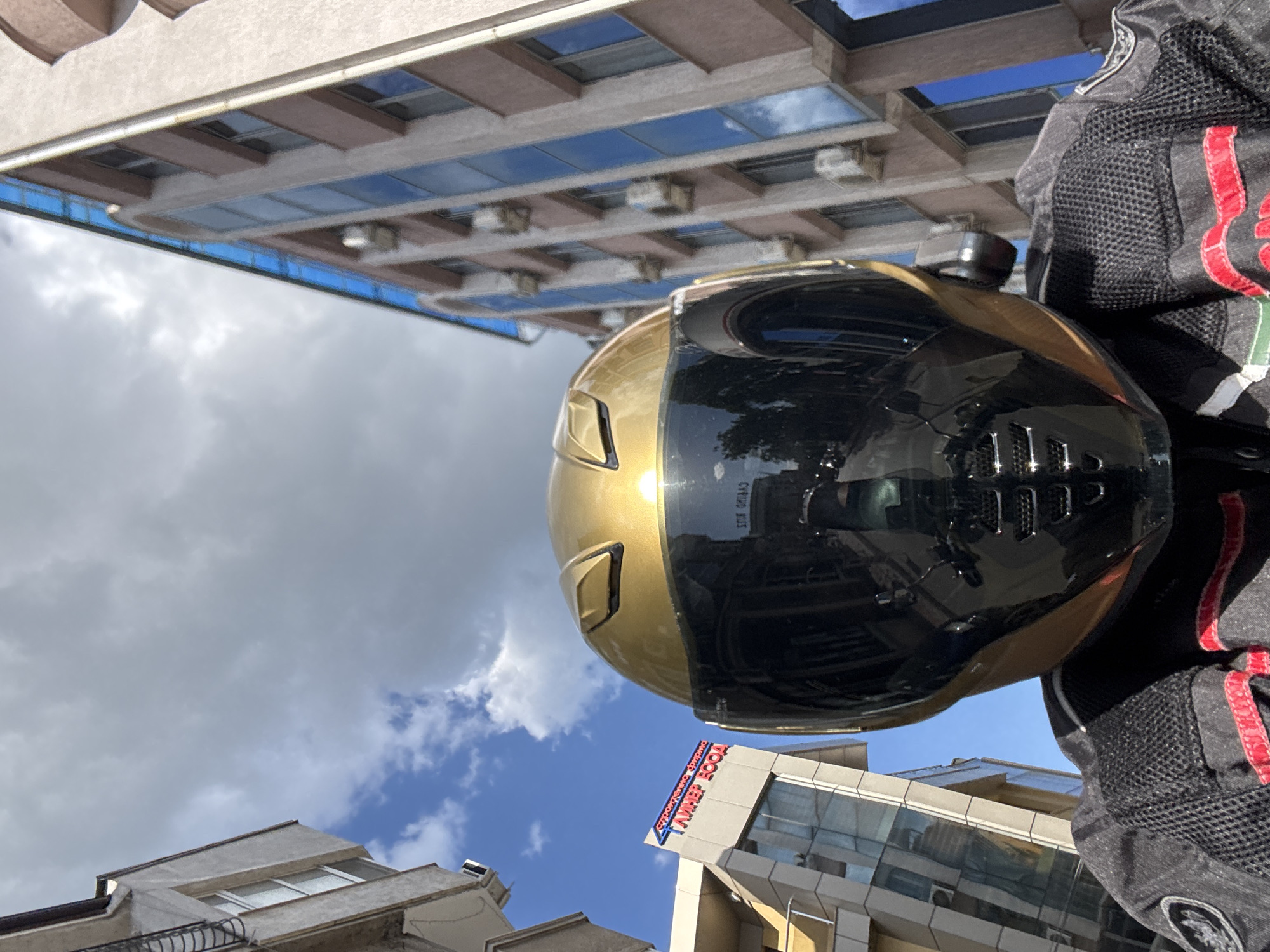 The image shows a low-angle view with a motorcycle helmet and rider's jacket as the primary subjects. The helmet is gold with a black visor, reflecting the surroundings. The rider's jacket is black mesh with red accents.
The background includes buildings with varied architectural styles and glass facades. The sky is visible with a mix of blue and cloudy areas. A sign with Cyrillic text is visible on one of the buildings.
Location: Unknown, Unknown. It is not possible to identify the location based on the visual information in the image.