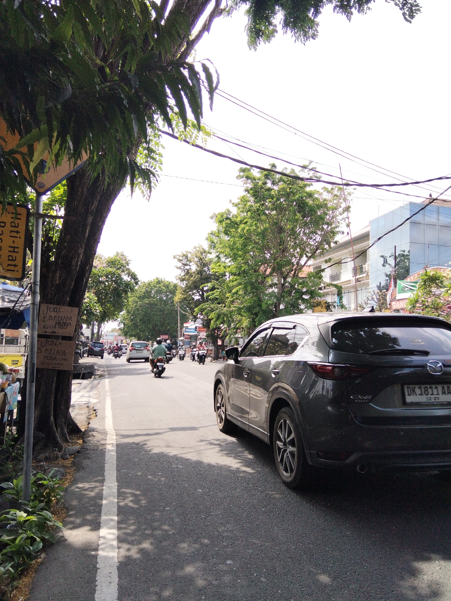 The image depicts a street scene in Denpasar, Indonesia, likely on a sunny day. In the foreground, a dark gray Mazda CX-5 is parked on the right side of the road, facing away from the viewer. Its license plate reads "DK 1811 AA" with additional smaller text below. The car's taillights and wheels are visible.
To the left of the car, a paved road runs parallel to a sidewalk. A large tree with dense foliage shades a section of the sidewalk and the road. Attached to the tree is a pole with several signs. One sign is yellow with black text, and another is a wooden sign with handwritten text in Indonesian. The text on the wooden sign appears to be an advertisement for selling coconuts ("JUAL ES KELAPA MUDA GULA ASLI" which translates to "SELLING YOUNG COCONUT ICE WITH ORIGINAL SUGAR").
Further down the road, several motorcycles and a white car are traveling in the same direction as the parked Mazda. The road is lined with trees and buildings on both sides. One building on the right is modern with a glass facade. The sky is bright and overcast. The scene suggests a typical urban street environment in Denpasar, characterized by traffic, street vendors, and lush vegetation.