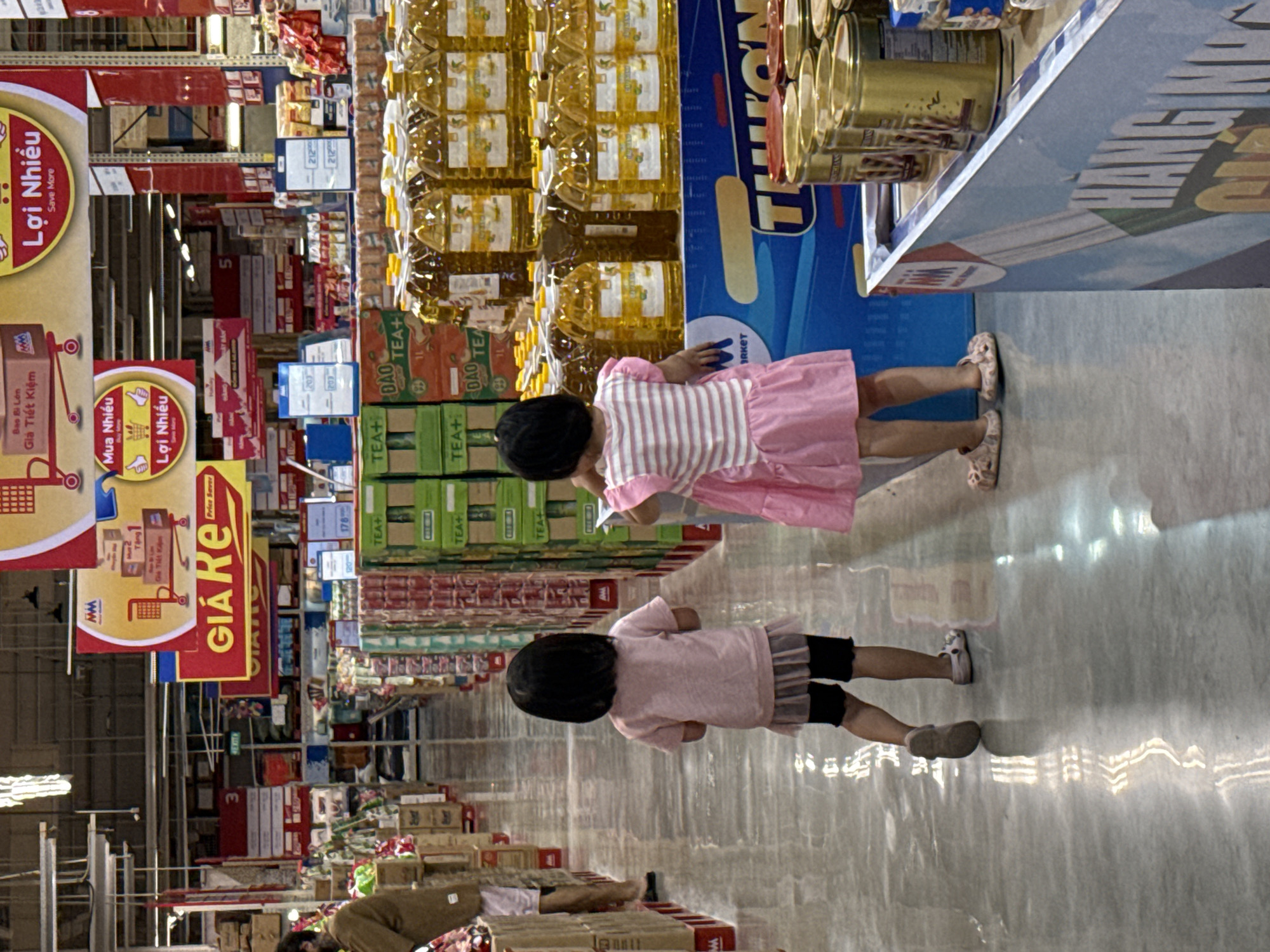 Two young girls are standing in the aisle of a large retail store, possibly in Tay Nha Trang Ward, Vietnam. They appear to be looking at merchandise. One girl is wearing a pink and white striped top and pink skirt, and the other is wearing a light pink top and black shorts. The aisle is stocked with various items, including bottled cooking oil and tea. Above the aisles are promotional signs written in Vietnamese. The overall setting is a busy commercial environment, suggesting a local supermarket or hypermarket.