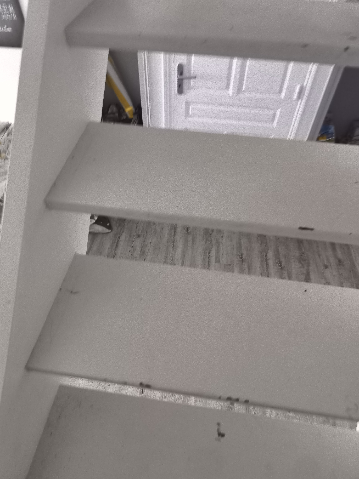 The image depicts a close-up, upward view of a white staircase. Through the risers, the scene reveals a glimpse of a room with a white door featuring a horizontal rectangular pattern and a silver-colored handle. A section of flooring with a wood-like pattern is also visible. At least one item with a yellow component is visible behind the stairs. The photograph does not contain any recognizable information to specify the location as Pélussin, France, or any other specific place. The image does not depict any people, subjects, or actions.