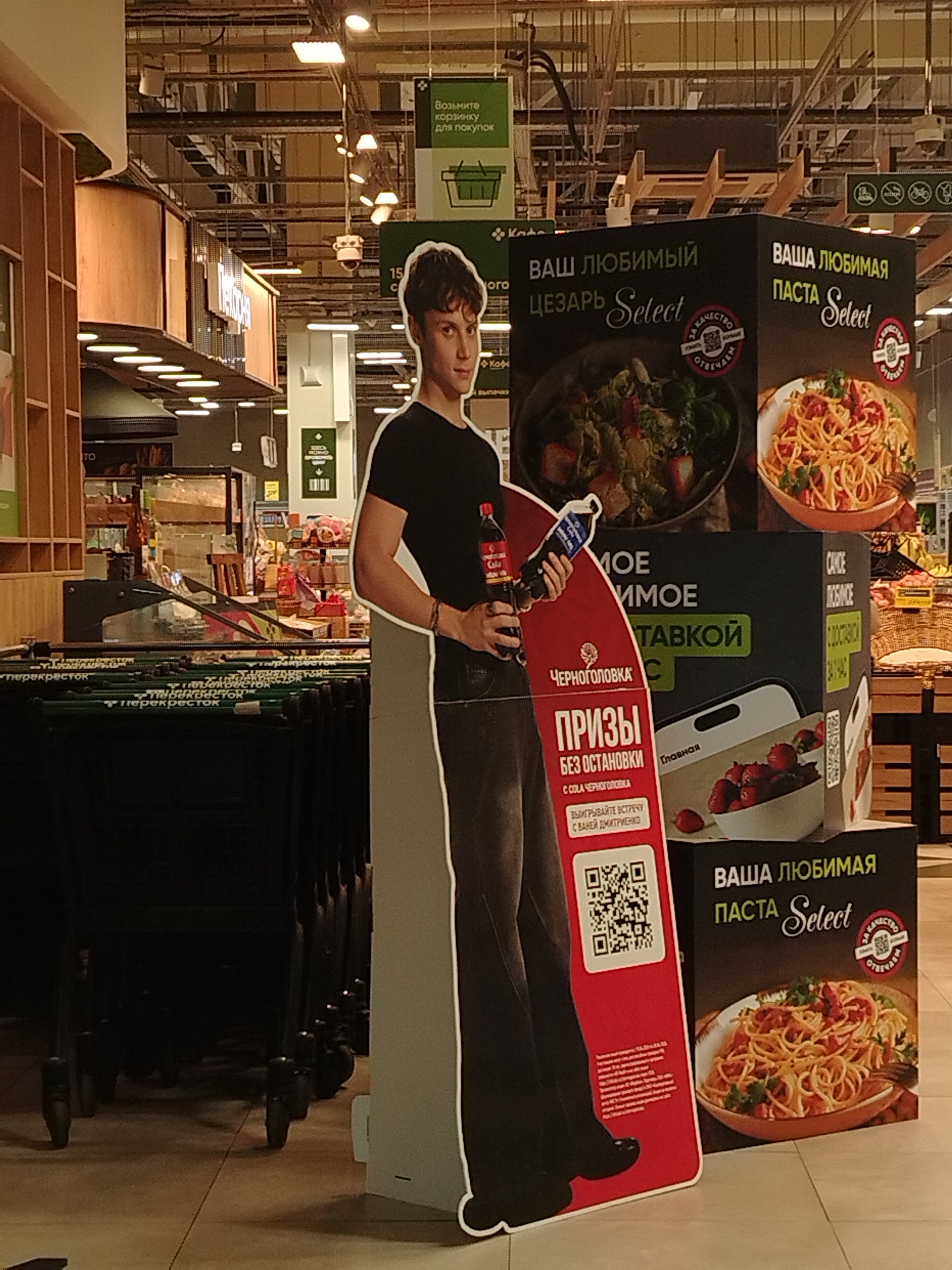 The image shows the interior of a supermarket, presumably in Izhevsk, Russia. There is a cardboard cutout of a young man holding two bottles of "Chernogolovka" cola, a promotional display for the drink. Beside the cutout are stacked boxes advertising "Vasha Lyubimaya Pasta Select" and "Vash Lyubimiy Cezar Select." Shopping carts are visible in the foreground. Above, hanging signs direct customers, one stating "Take a basket for shopping" in Russian. The supermarket appears to be well-lit with many other displays, products, and shoppers visible in the background.