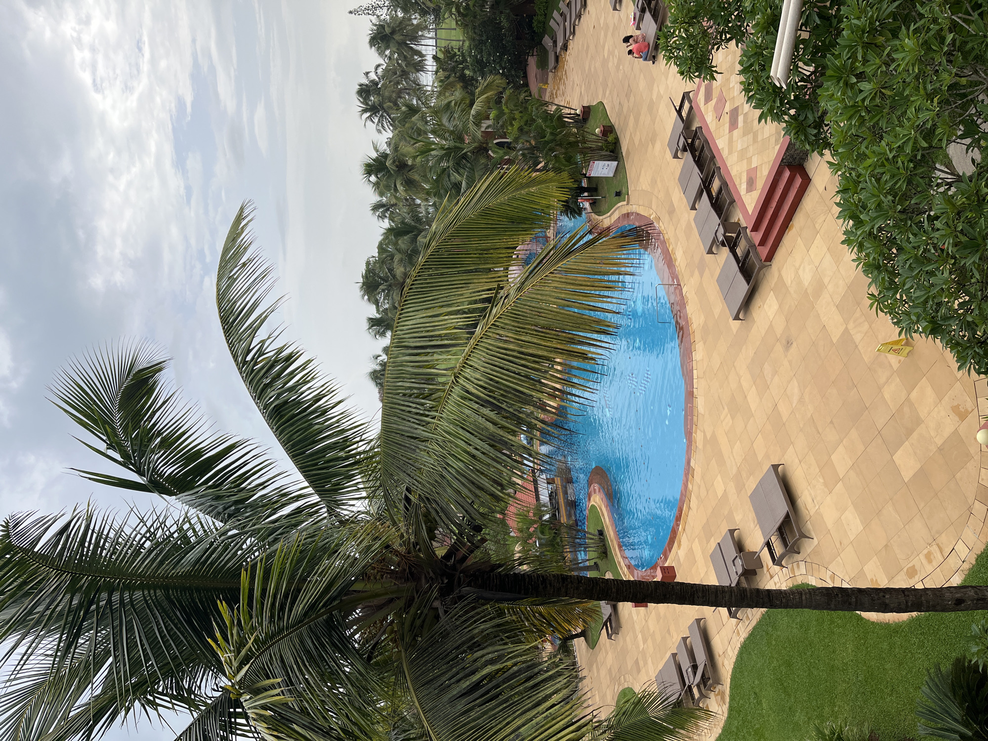 The image shows a resort pool area in Fatrade, India, on a cloudy day. A large, bright blue swimming pool with a curved shape is the central feature. It is surrounded by a paved area made of light brown square tiles. Several lounge chairs are arranged on the paving stones, some of which are empty and others occupied.
In the foreground, a tall palm tree with lush green fronds dominates the left side of the frame, its trunk a dark brown. On the right side of the pool, a group of people is visible, seated on lounge chairs.
Beyond the pool and paved area, there are more palm trees and other tropical foliage, suggesting a resort or hotel setting. In the background, a glimpse of buildings with red tiled roofs can be seen, as well as more distant trees and a hint of the sky above, characterized by overcast clouds. The overall scene depicts a serene and tropical environment.