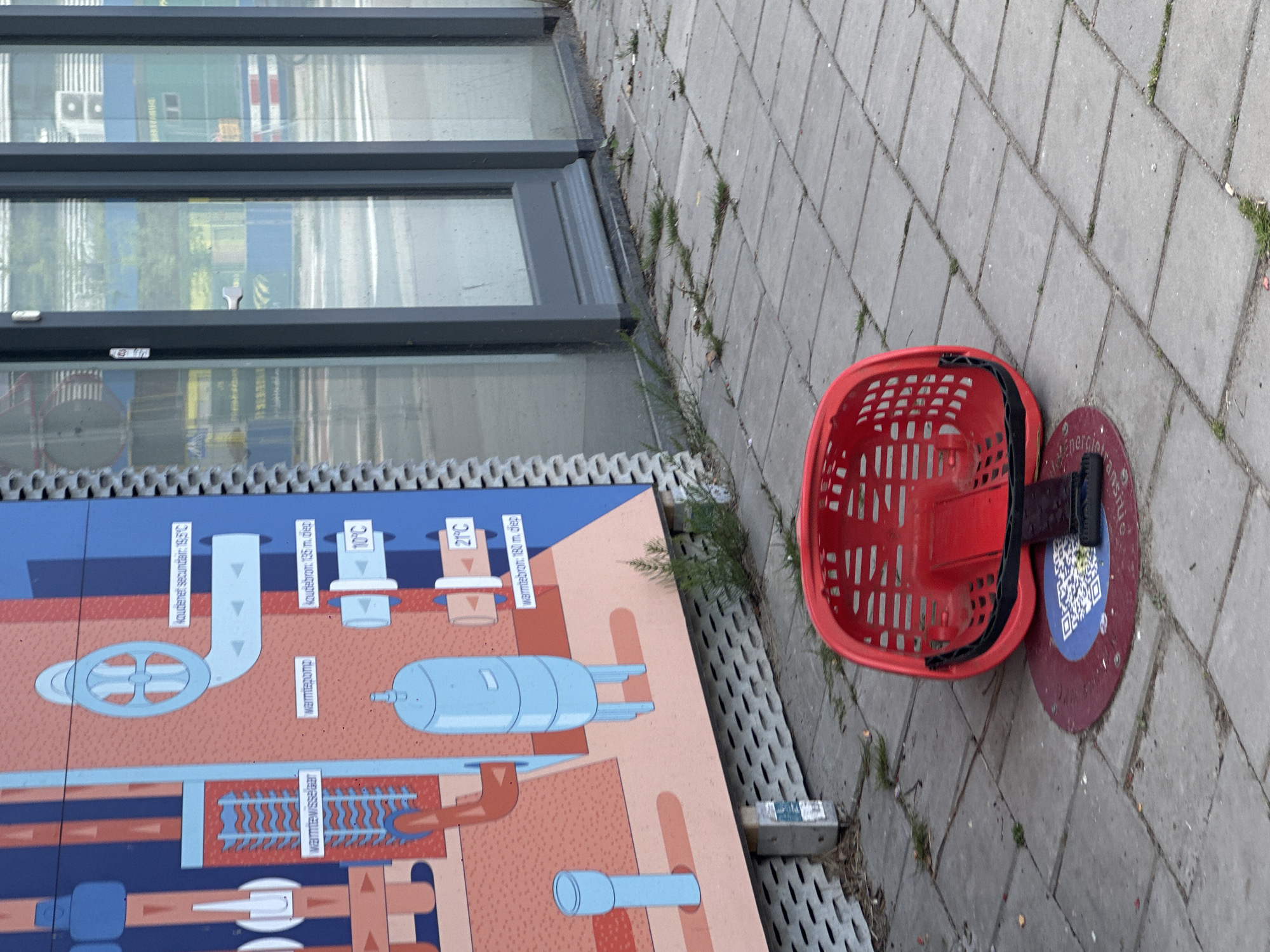 The image shows an outdoor urban scene likely in Amsterdam, Netherlands. The foreground features a red plastic shopping basket attached to a dark-colored bike-sharing stand. The stand is circular with a QR code on it. The basket and stand rest on a paved surface with some grass growing in the cracks. To the left, there's a diagram illustrating a geothermal energy system with text labels in Dutch, including "Koudebron 135 m diep", "warmtebron 180 m diep", and "Warmtewisselaar". The diagram is mounted next to a building with large windows that reflect the surrounding environment.