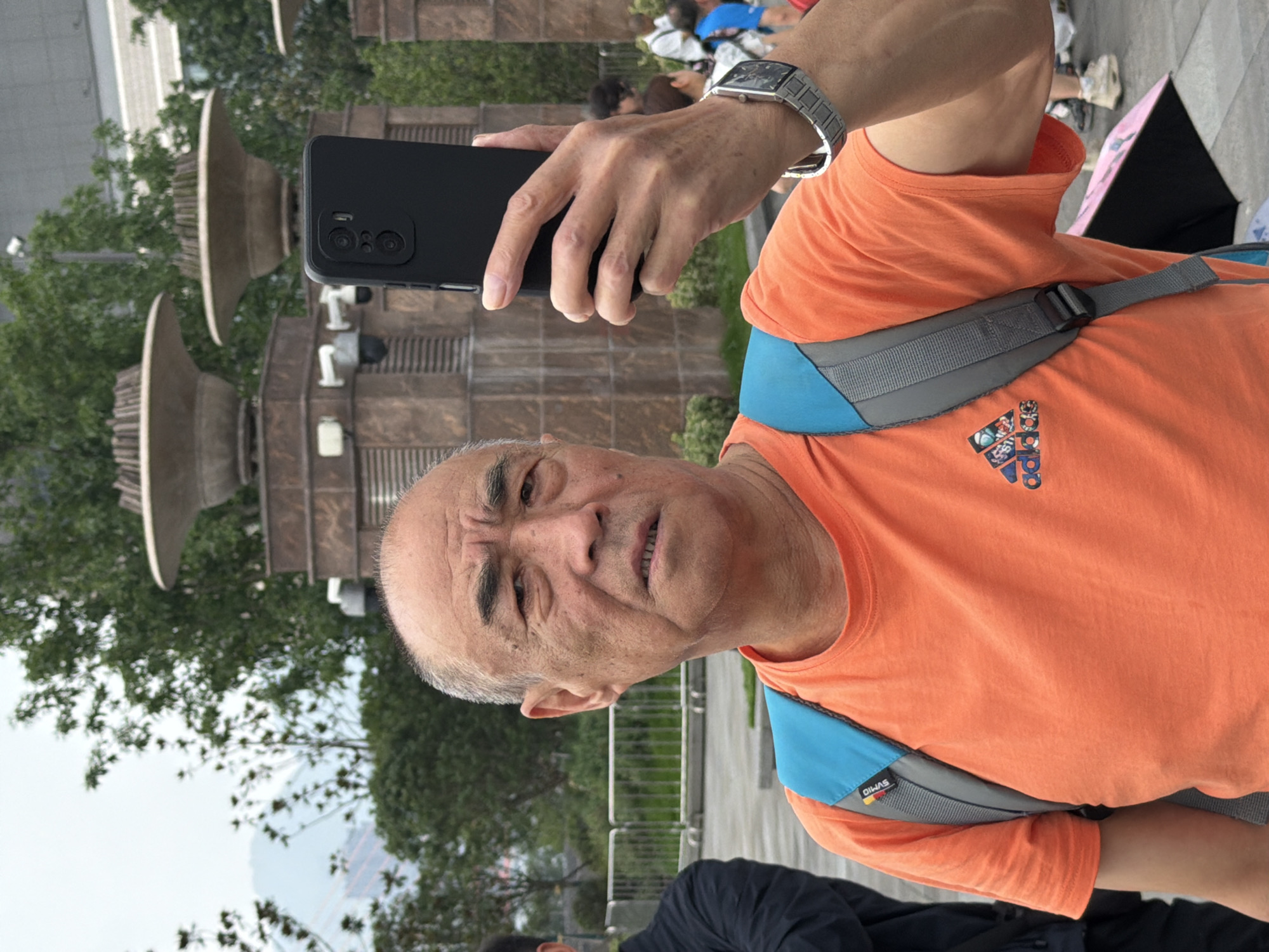 An older East Asian man in an orange t-shirt and backpack stands in the foreground, holding a black smartphone up to take a picture of himself. The man has short gray hair and appears to be looking at the phone's screen. He is wearing a silver digital watch on his left wrist. Behind him, a modern urban environment is visible with trees, buildings, and surveillance cameras mounted on a stone structure. In the background, people are walking and blurred out. The scene appears to be outdoors in a public space within Yuzhong District, China, possibly a park or plaza, indicated by the presence of trees, urban architecture, and a paved walkway.
