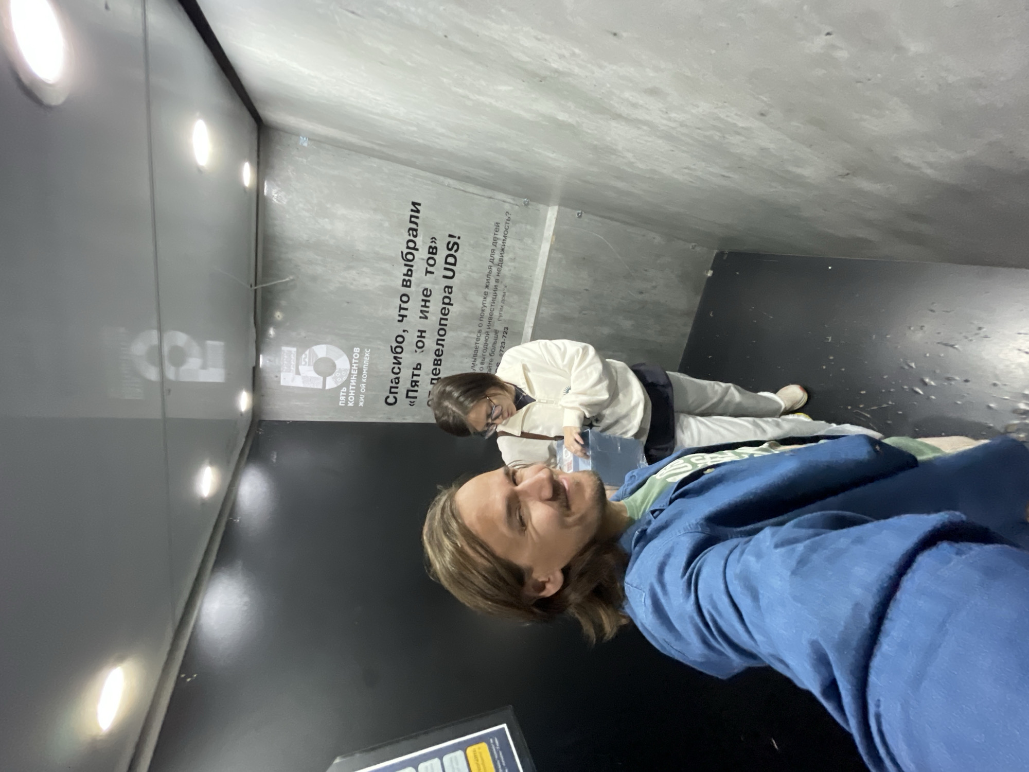 A man is taking a selfie with another woman in a building. They are standing in a concrete room with "Пять континентов" on the wall and Russian text. The man has light brown hair and is wearing a blue shirt. The woman is wearing glasses and a white jacket. They are both holding a book.